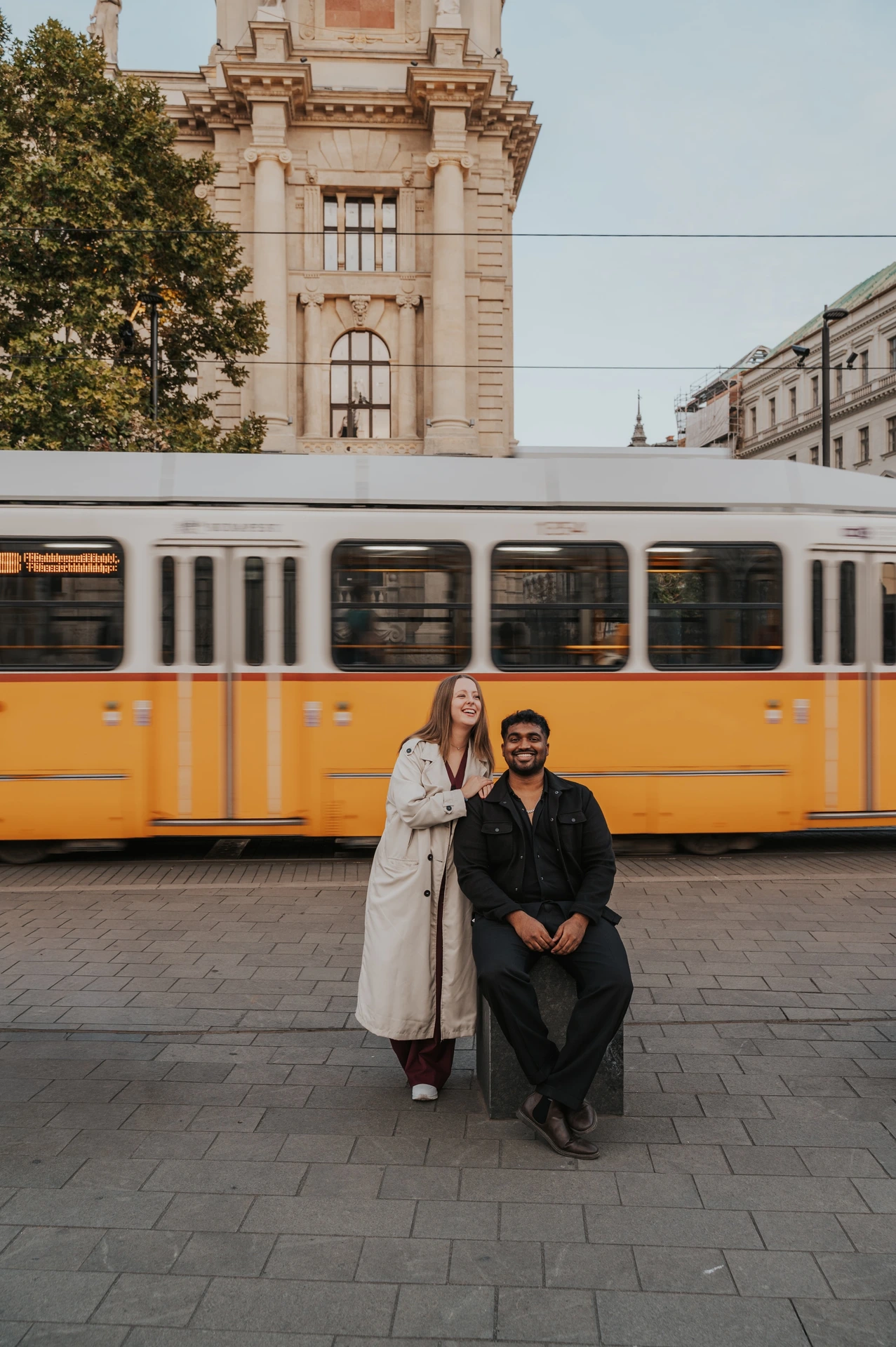 Budapest couple photoshoot: a yellow tram behind their city portrait The couple posing beside a stone bollard, with her in a light coat and a yellow Budapest tram passing behind them during their Budapest couple photoshoot.