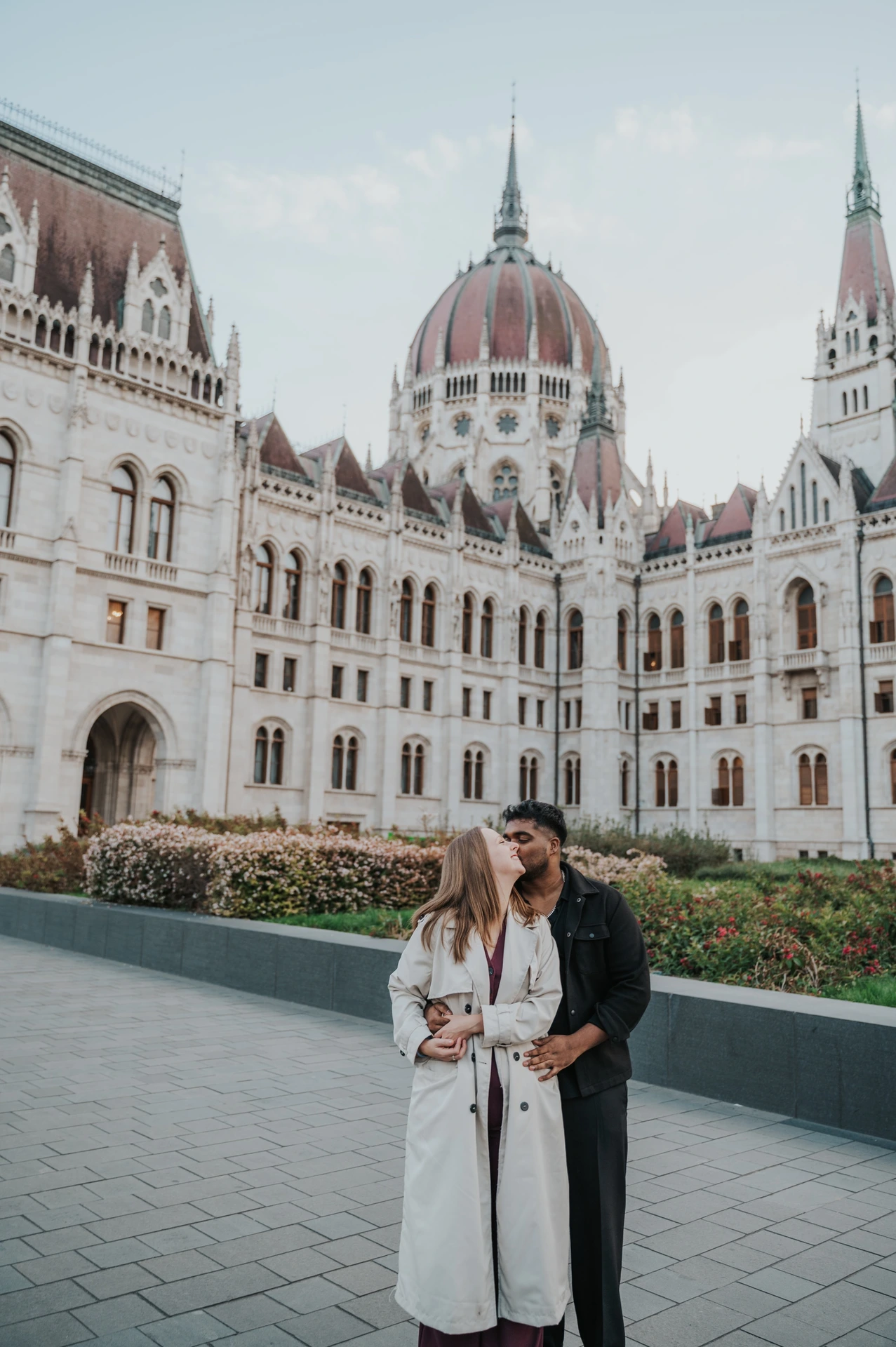 Budapest couple photoshoot: an embrace framed by Parliament’s towers A full-length view of the couple embracing beside the garden wall, with Parliament’s dome and pointed towers around them during their Budapest couple photoshoot.