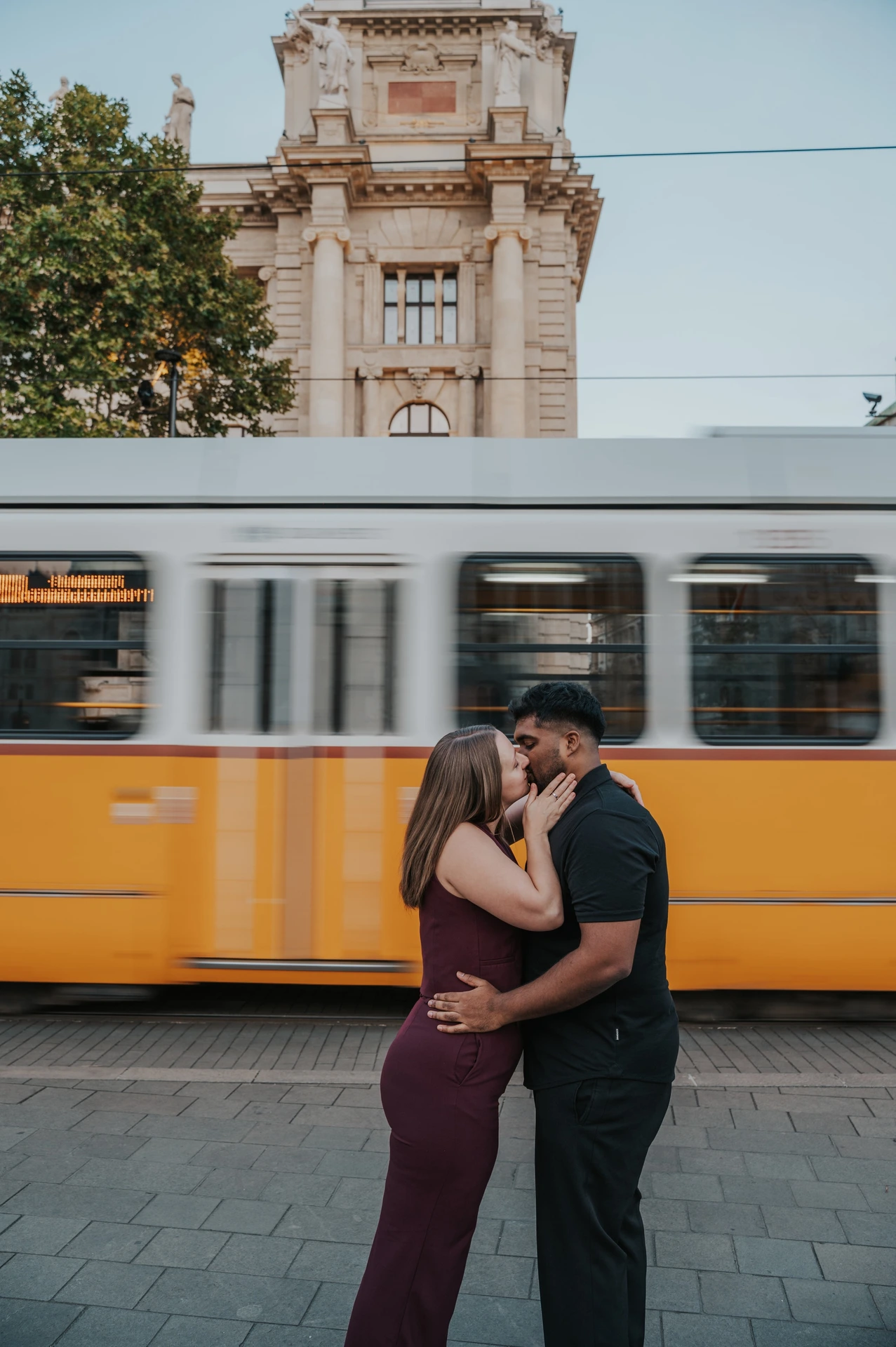 Budapest couple photoshoot: an embrace with the tram passing behind The couple embracing and kissing beside the tram route, with the yellow tram’s windows blurred behind them during their Budapest couple photoshoot.