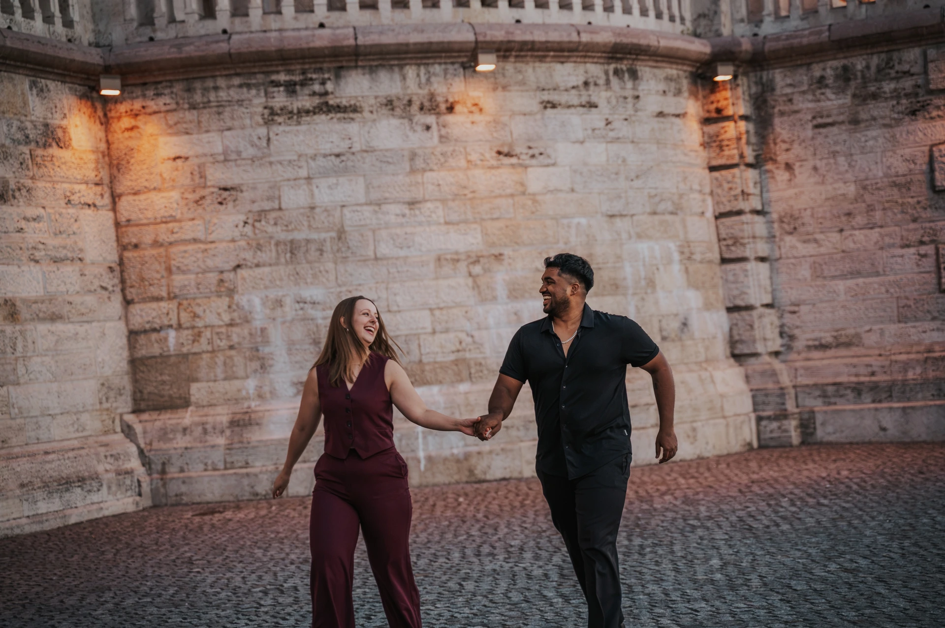 Budapest couple photoshoot: an evening walk beside Parliament’s stone wall A landscape photograph of the couple walking hand in hand beside Parliament’s stone wall, with warm lamps lighting the background during their Budapest couple photoshoot.