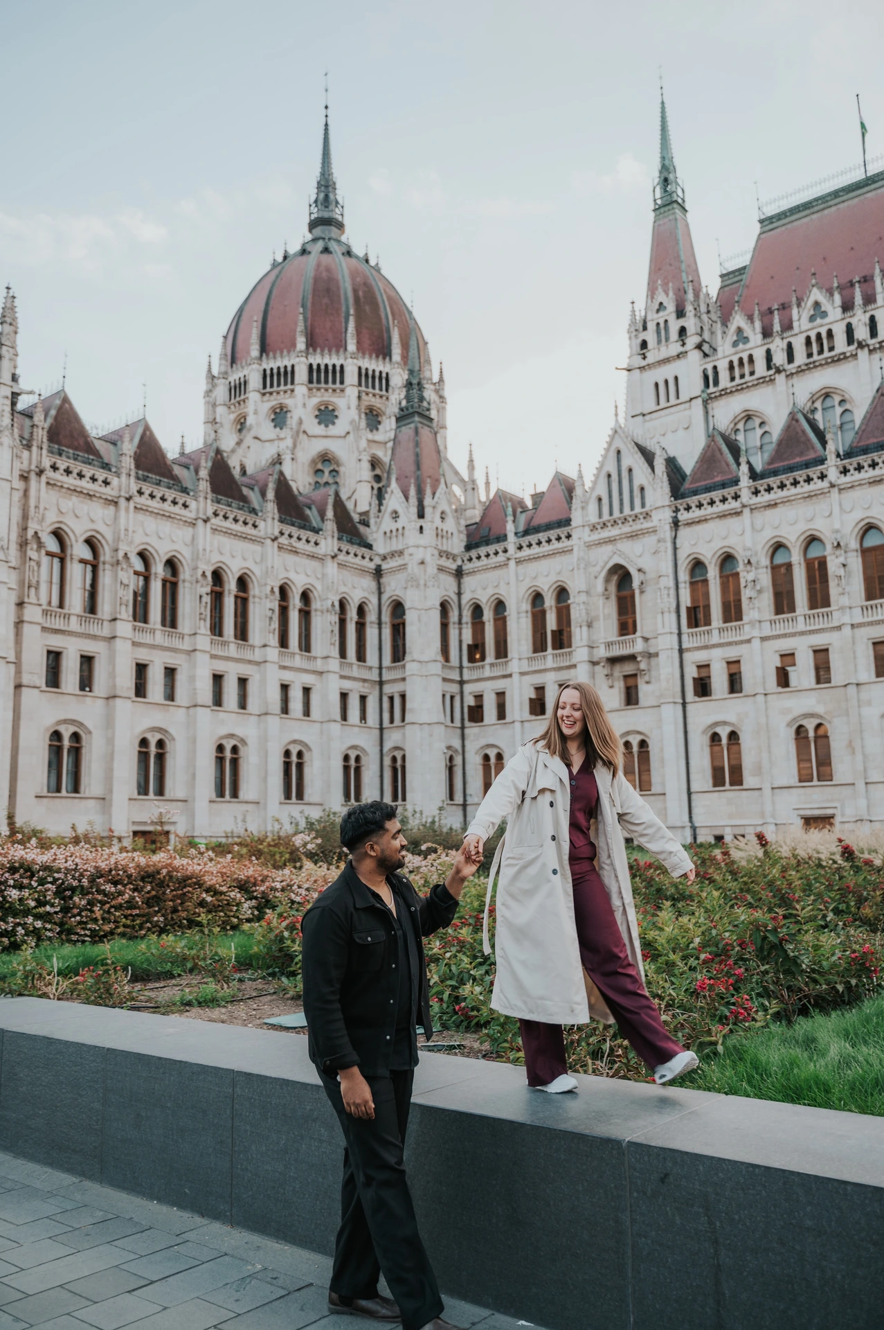 She balances along the low garden ledge while holding his hand, with the Hungarian Parliament and flowerbeds in the background during their Budapest couple photoshoot.