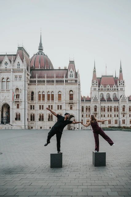 The couple holding hands while balancing on separate low stone bollards, each stretching out a leg in Parliament Square during their Budapest couple photoshoot.