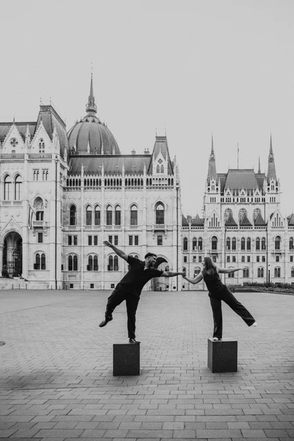 Budapest couple photoshoot: black-and-white balance and laughter Black-and-white photograph of the couple balancing on two low stone bollards and holding hands, with Parliament behind them during their Budapest couple photoshoot.