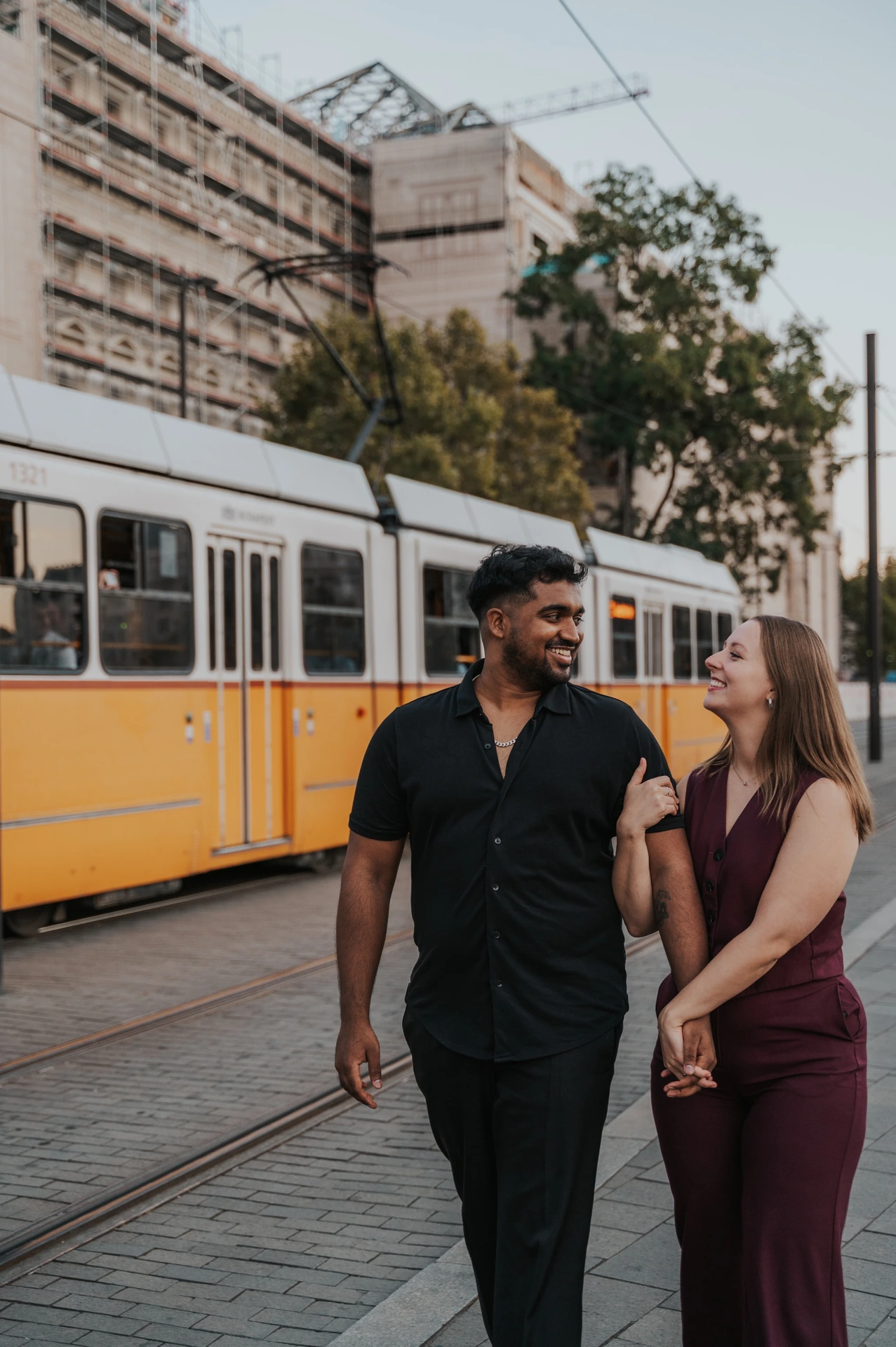 Budapest couple photoshoot: close smiles beside a characteristic yellow tram A closer photograph of the couple smiling at one another as they walk hand in hand, with a yellow Budapest tram behind them during their Budapest couple photoshoot.