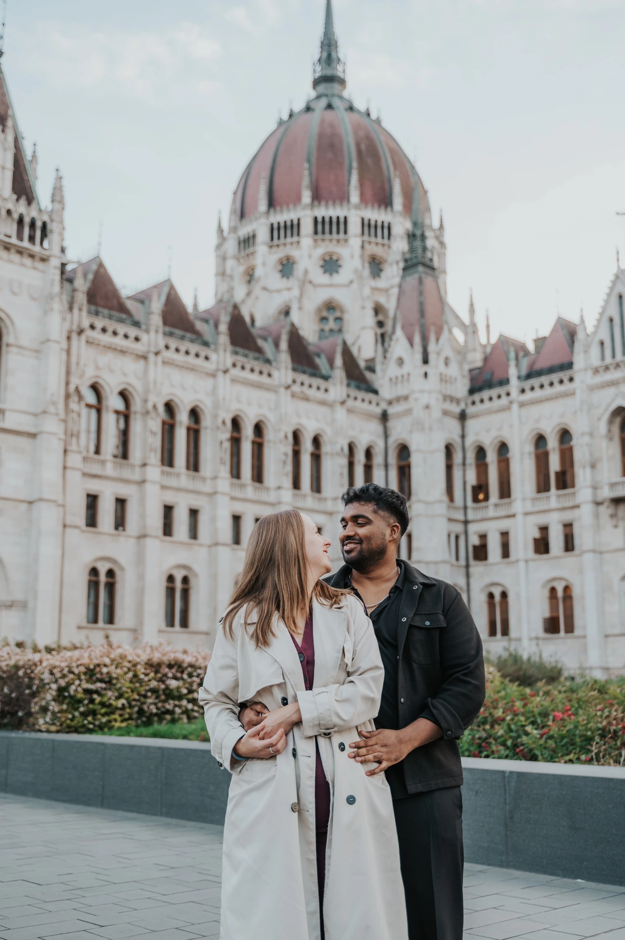 The couple smiling towards one another, his arm around her waist, with Parliament and the flowerbeds behind them during their Budapest couple photoshoot.