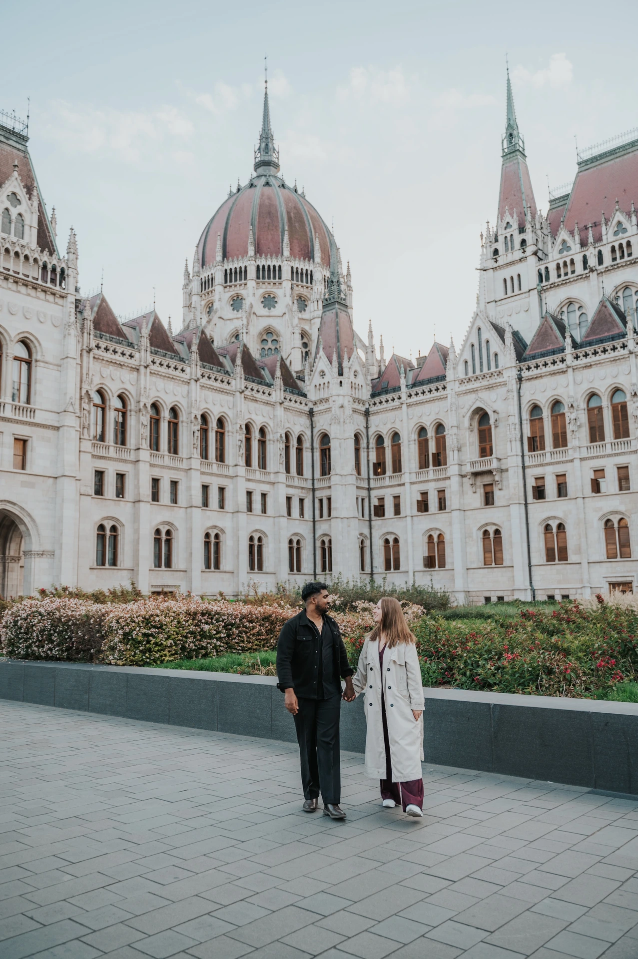 Budapest couple photoshoot: hand in hand beside Parliament’s flowerbeds The couple holding hands beside the low garden wall, with Parliament’s pale stone towers and dome filling the background during their Budapest couple photoshoot.