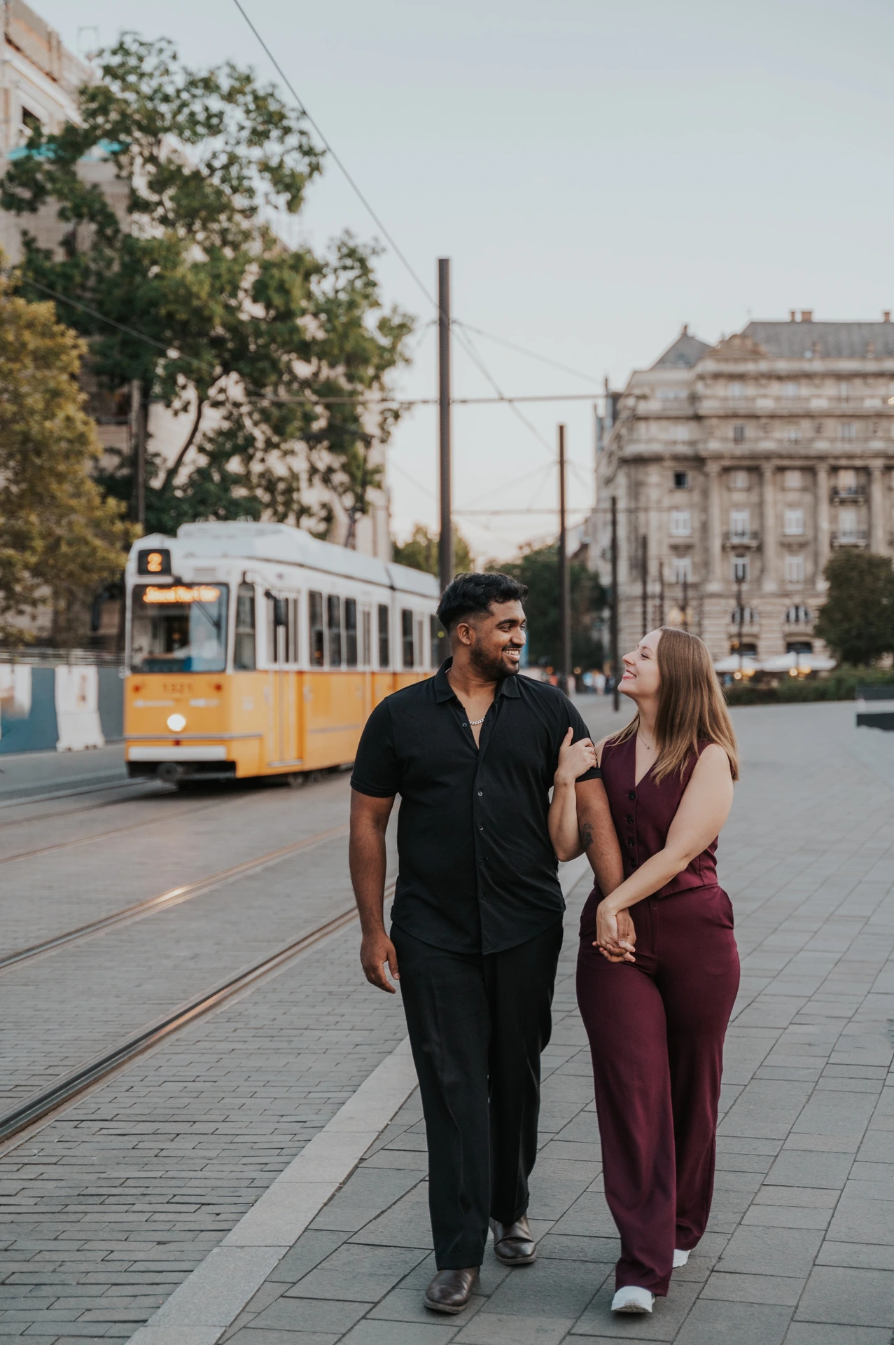 Budapest couple photoshoot: hand in hand with a tram behind them The couple holding hands and smiling at one another while walking, with a yellow tram, trees and city buildings behind them during their Budapest couple photoshoot.