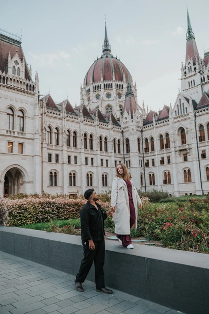 Budapest couple photoshoot: holding hands at different heights She stands on the low garden ledge while he stands below, the couple holding hands with Parliament behind them during their Budapest couple photoshoot.