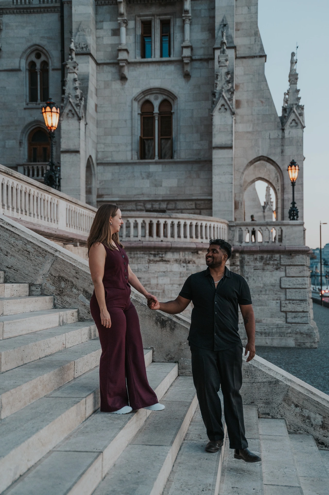 She stands one step above him as the couple hold hands on Parliament’s staircase, with carved arches and a glowing lamp behind them during their Budapest couple photoshoot.