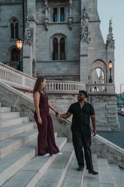 She stands one step above him as the couple hold hands on Parliament’s staircase, with carved arches and a glowing lamp behind them during their Budapest couple photoshoot.
