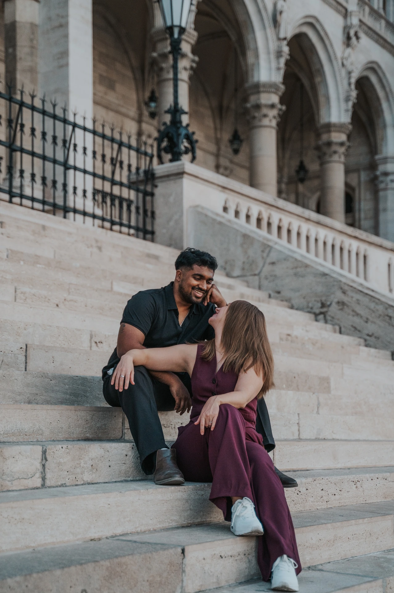 Budapest couple photoshoot: laughing together on the Parliament steps A closer view of the couple laughing together on Parliament’s stone steps, with her leaning towards him beneath the ornate arches during their Budapest couple photoshoot.
