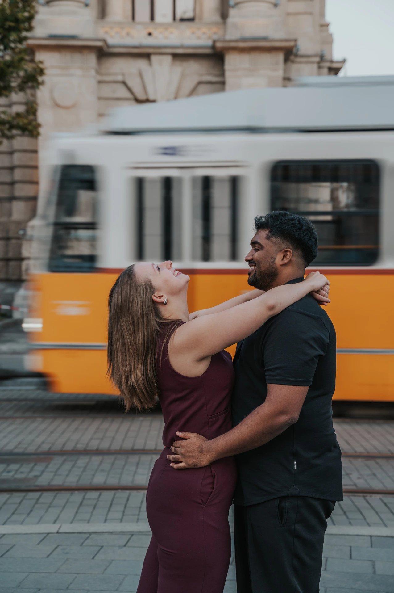 She leans back laughing while he holds her in an embrace, with a yellow Budapest tram passing behind them during their Budapest couple photoshoot.