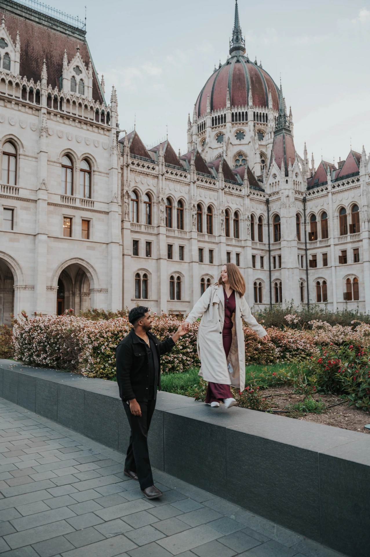 Budapest couple photoshoot: outstretched arms beside Parliament’s gardens She stretches out her free arm while balancing on the garden ledge and holding his hand beside Parliament’s flowerbeds during their Budapest couple photoshoot.