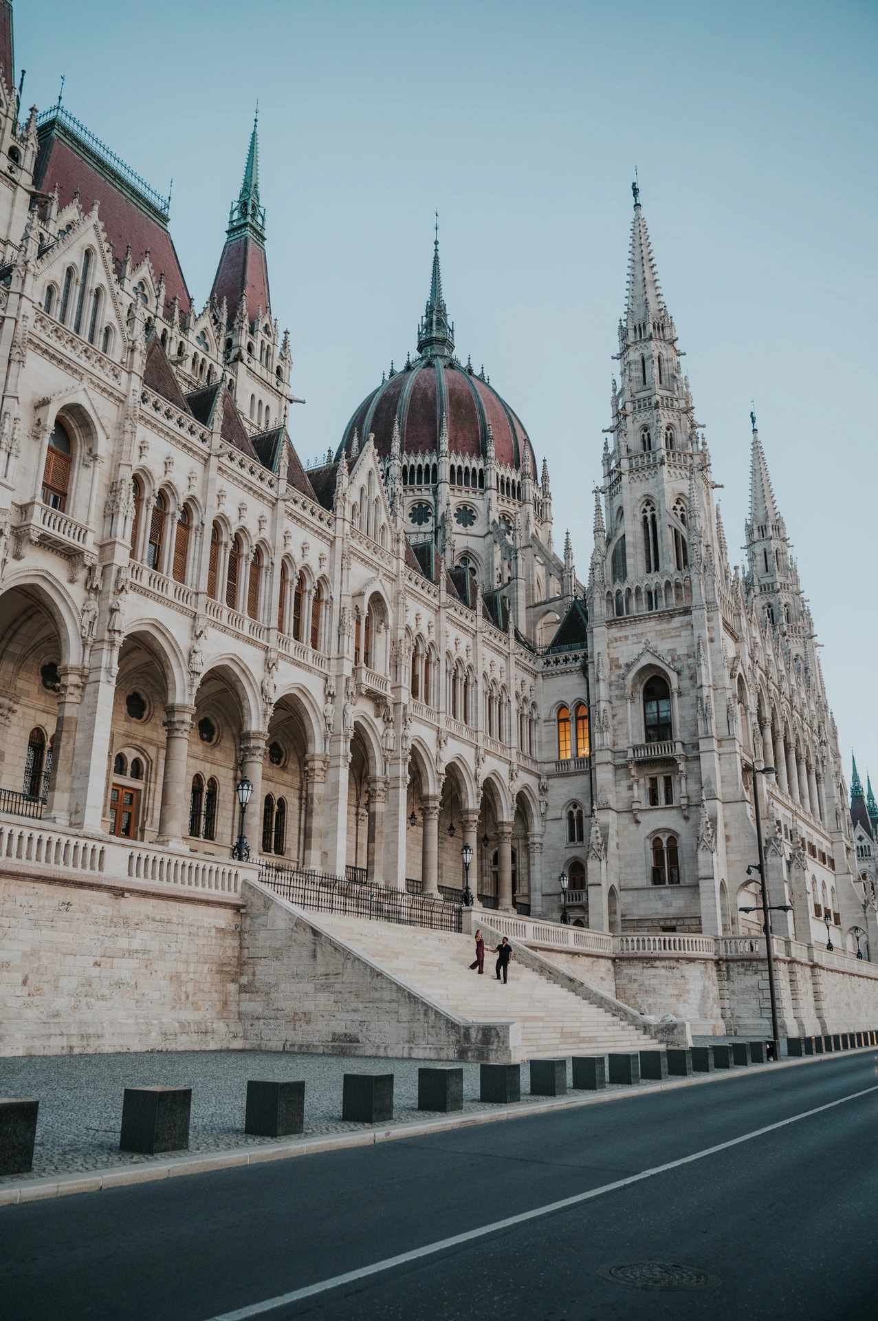 Budapest couple photoshoot: parliament’s riverside architecture at dusk An architectural view along the Hungarian Parliament’s ornate riverside façade, with stone steps, the road and the evening sky during their Budapest couple photoshoot.