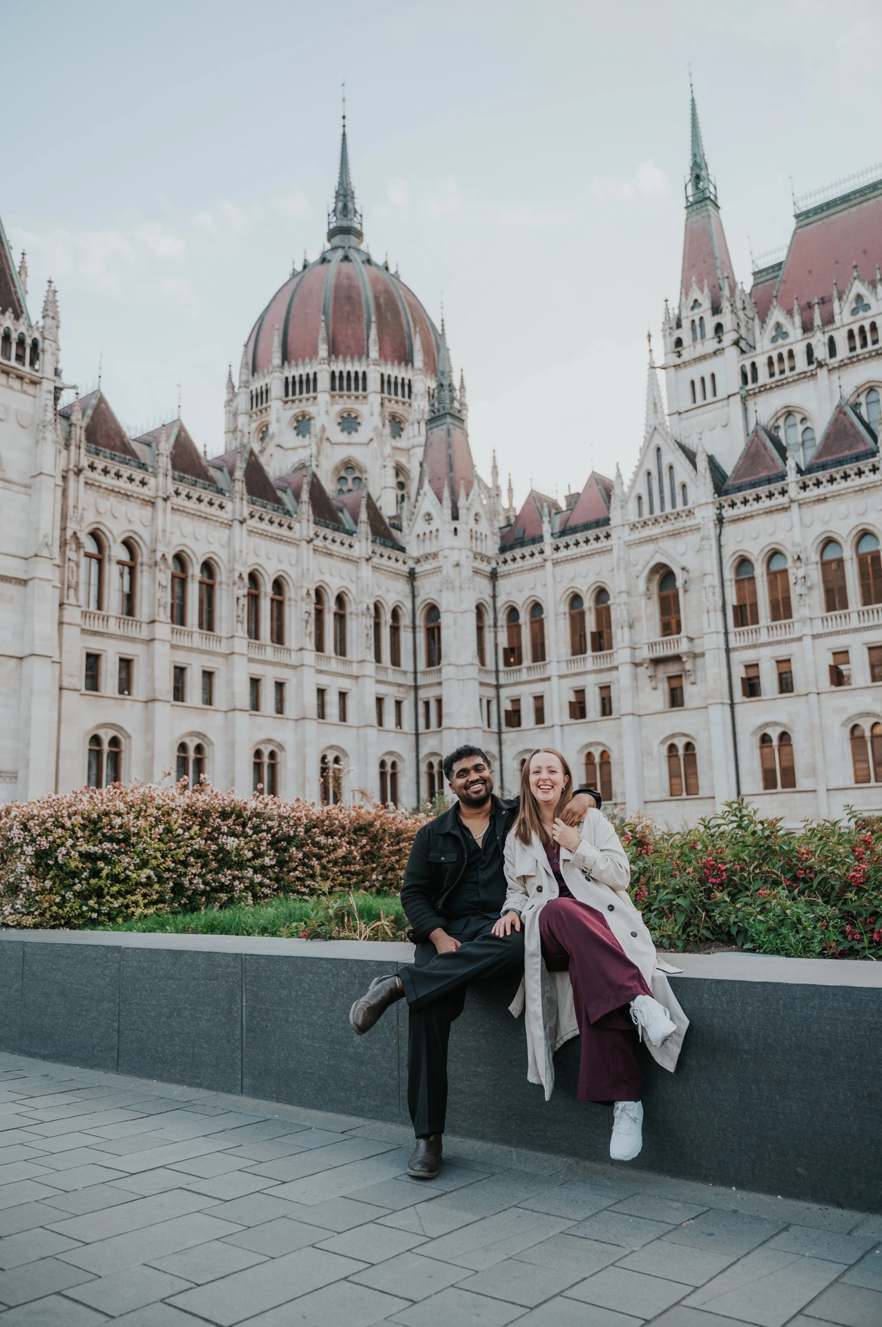 The couple raising their arms playfully while seated on the garden wall in front of the Hungarian Parliament during their Budapest couple photoshoot.