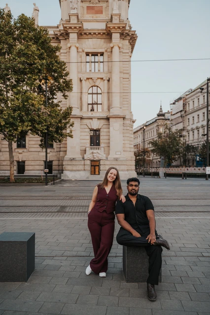 Budapest couple photoshoot: relaxed portraits beside the tram route The couple together by a stone bollard near the tram route, with historic Budapest buildings and a tree in the background during their Budapest couple photoshoot.