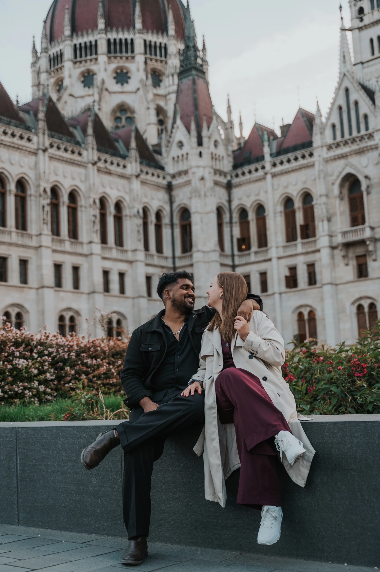 Budapest couple photoshoot: relaxed portraits on the garden ledge The couple looking towards one another while seated on the garden ledge outside the Hungarian Parliament during their Budapest couple photoshoot.