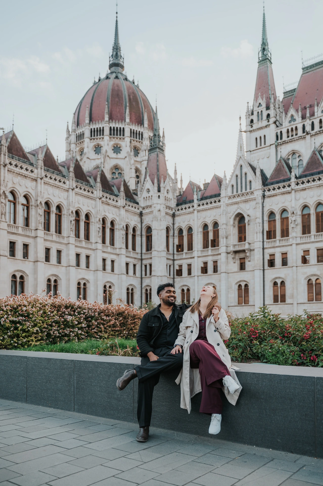 Budapest couple photoshoot: sharing smiles on the stone garden ledge The couple seated together and sharing a smile beside the flowerbeds, with Parliament’s dome in the backdrop during their Budapest couple photoshoot.