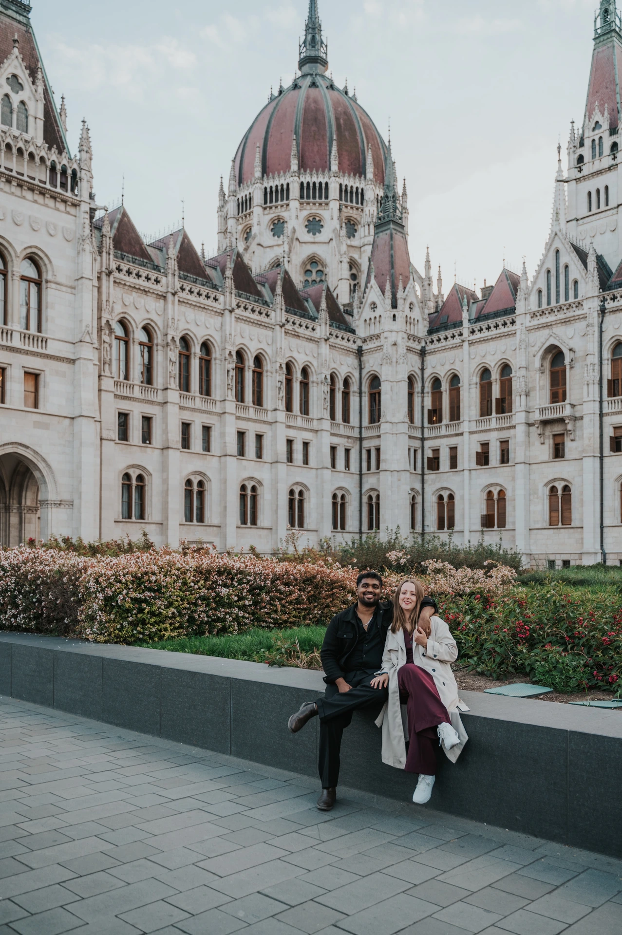 The couple sitting close together on the low garden wall, with the Hungarian Parliament and flowerbeds behind them during their Budapest couple photoshoot.