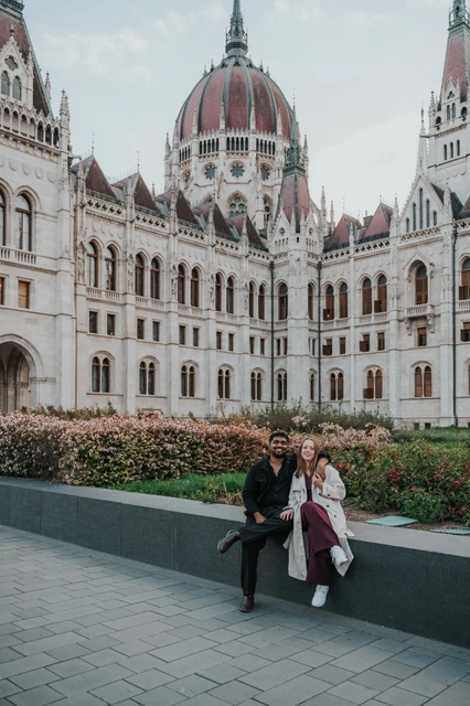Budapest couple photoshoot: sitting together on the Parliament garden wall The couple sitting close together on the low garden wall, with the Hungarian Parliament and flowerbeds behind them during their Budapest couple photoshoot.