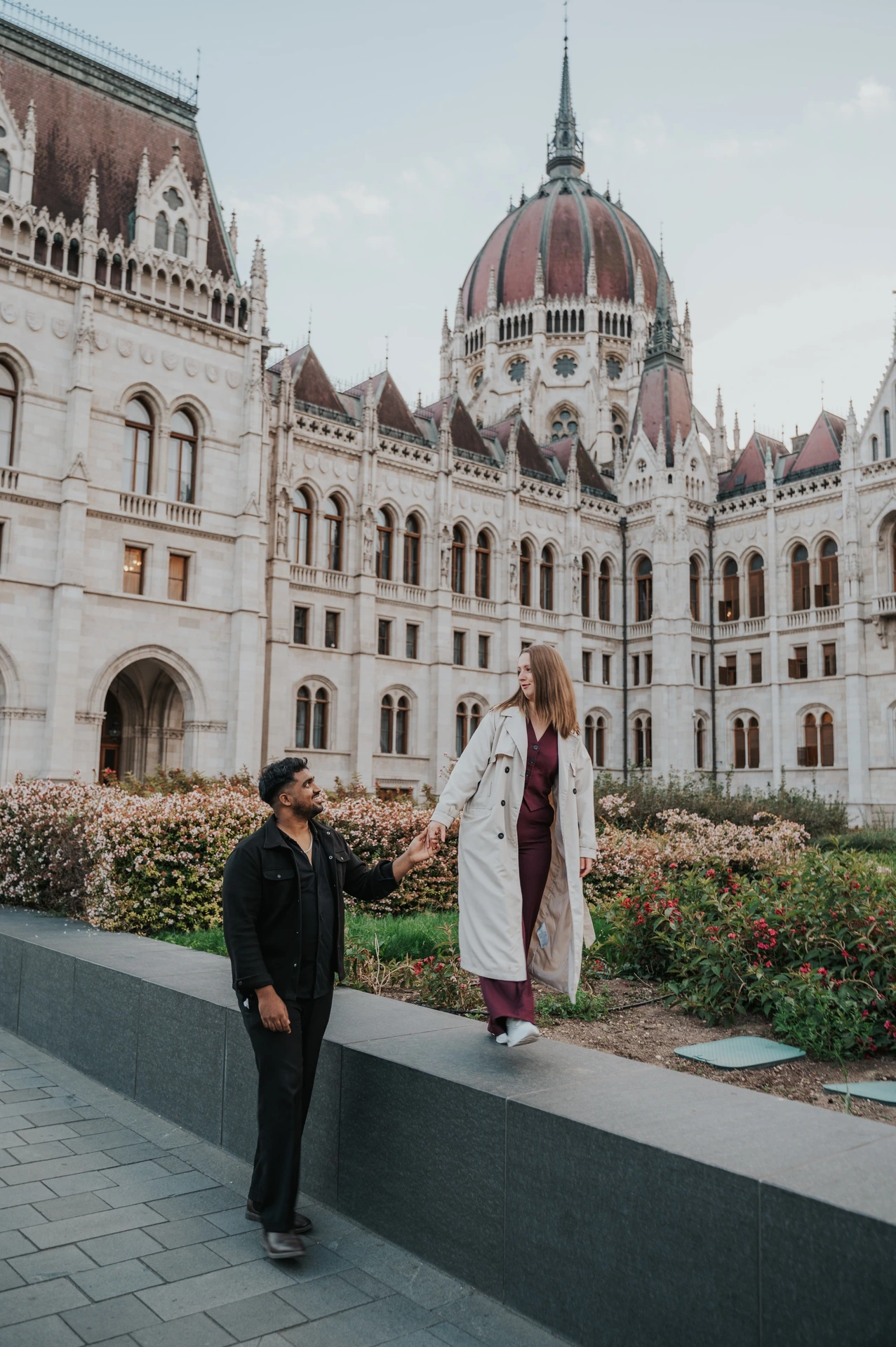The couple sharing a smile as she stands on the low garden ledge and he walks beside her outside Parliament during their Budapest couple photoshoot.