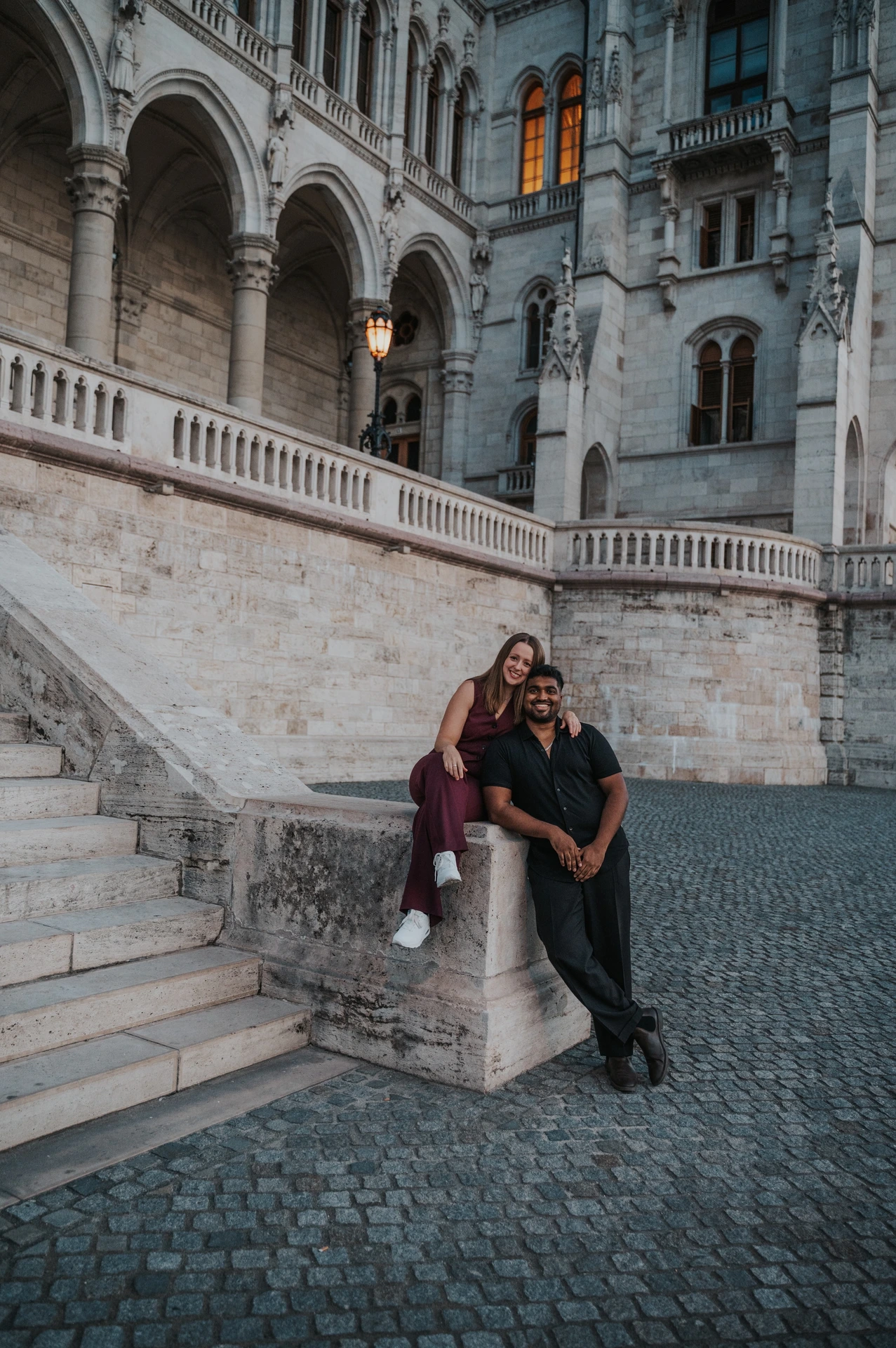 Budapest couple photoshoot: together beside the staircase’s stone pillar She sits on a low stone pillar beside Parliament’s staircase while he leans beside her, with carved arches and cobbled paving around them during their Budapest couple photoshoot.