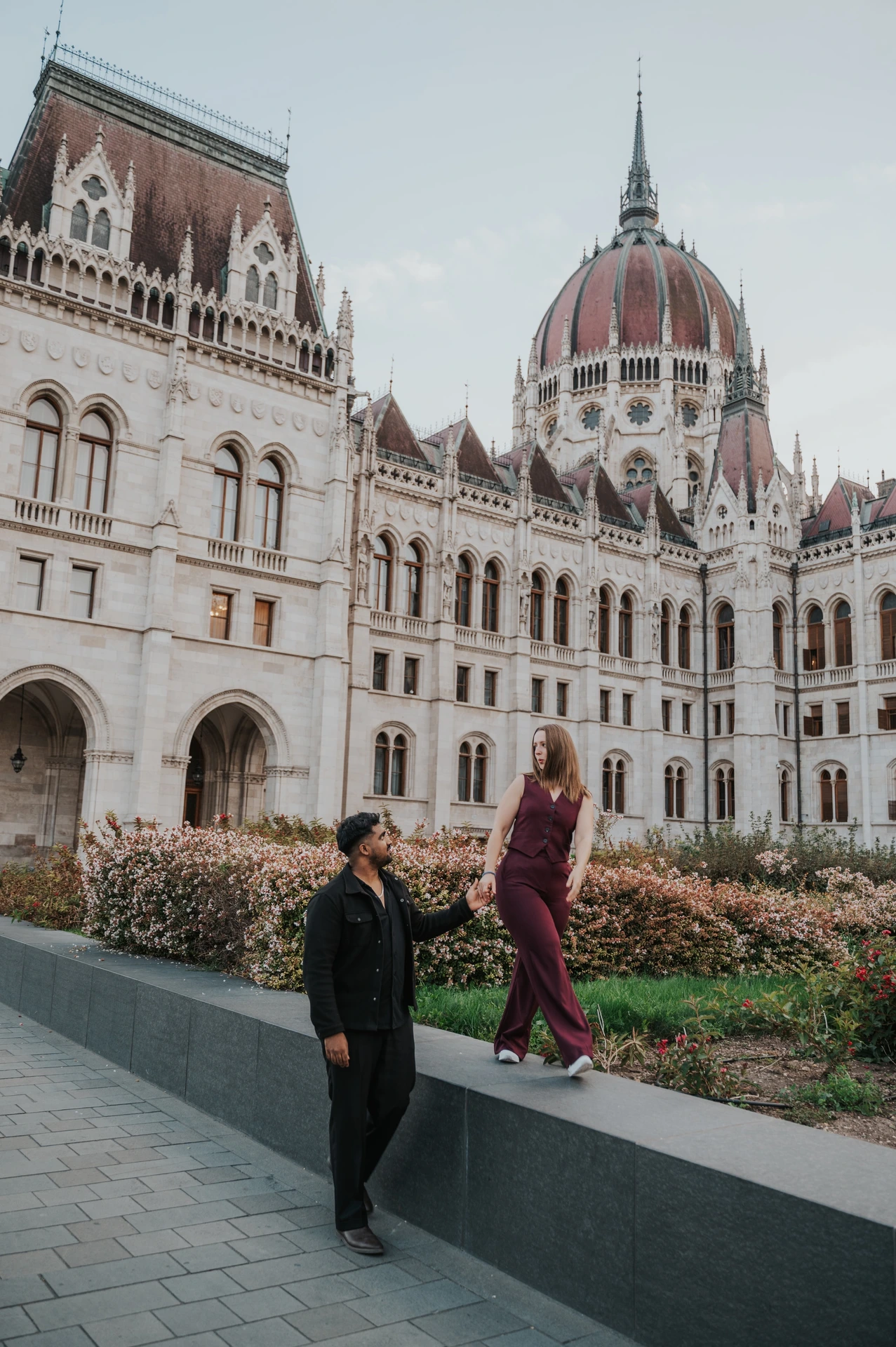Budapest couple photoshoot: walking hand in hand beside the flowerbeds The couple moving along the garden wall together, with her on the ledge and him on the paving beside Parliament during their Budapest couple photoshoot.