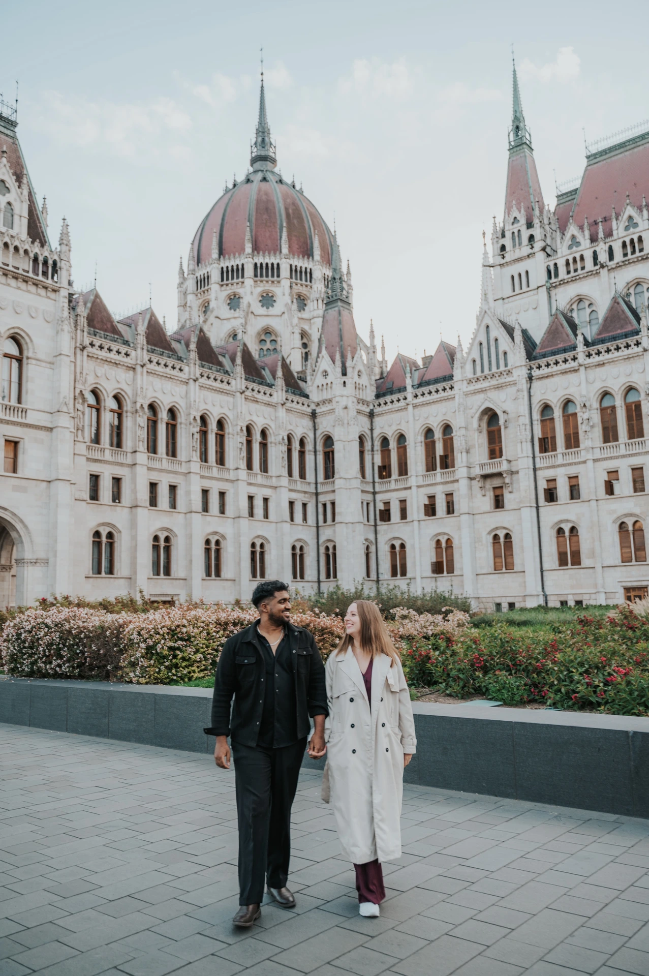 Budapest couple photoshoot: walking together beneath Parliament’s dome The couple walking together beside the flowerbeds, with the Hungarian Parliament’s dome and ornate façade behind them during their Budapest couple photoshoot.