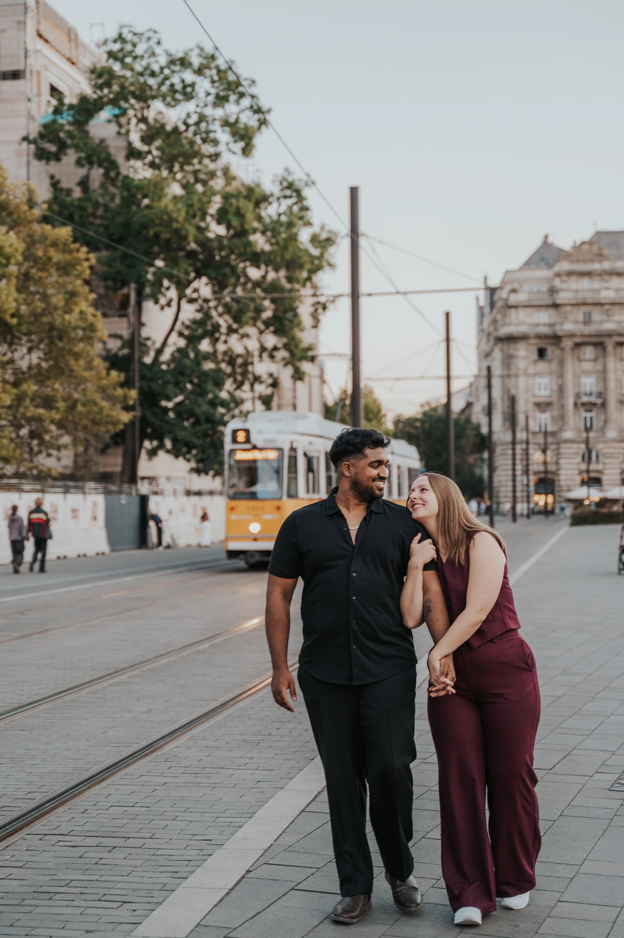 Budapest couple photoshoot: walking together beside the tram tracks The couple walking side by side and laughing near the tram tracks, with a yellow tram and historic buildings behind them during their Budapest couple photoshoot.