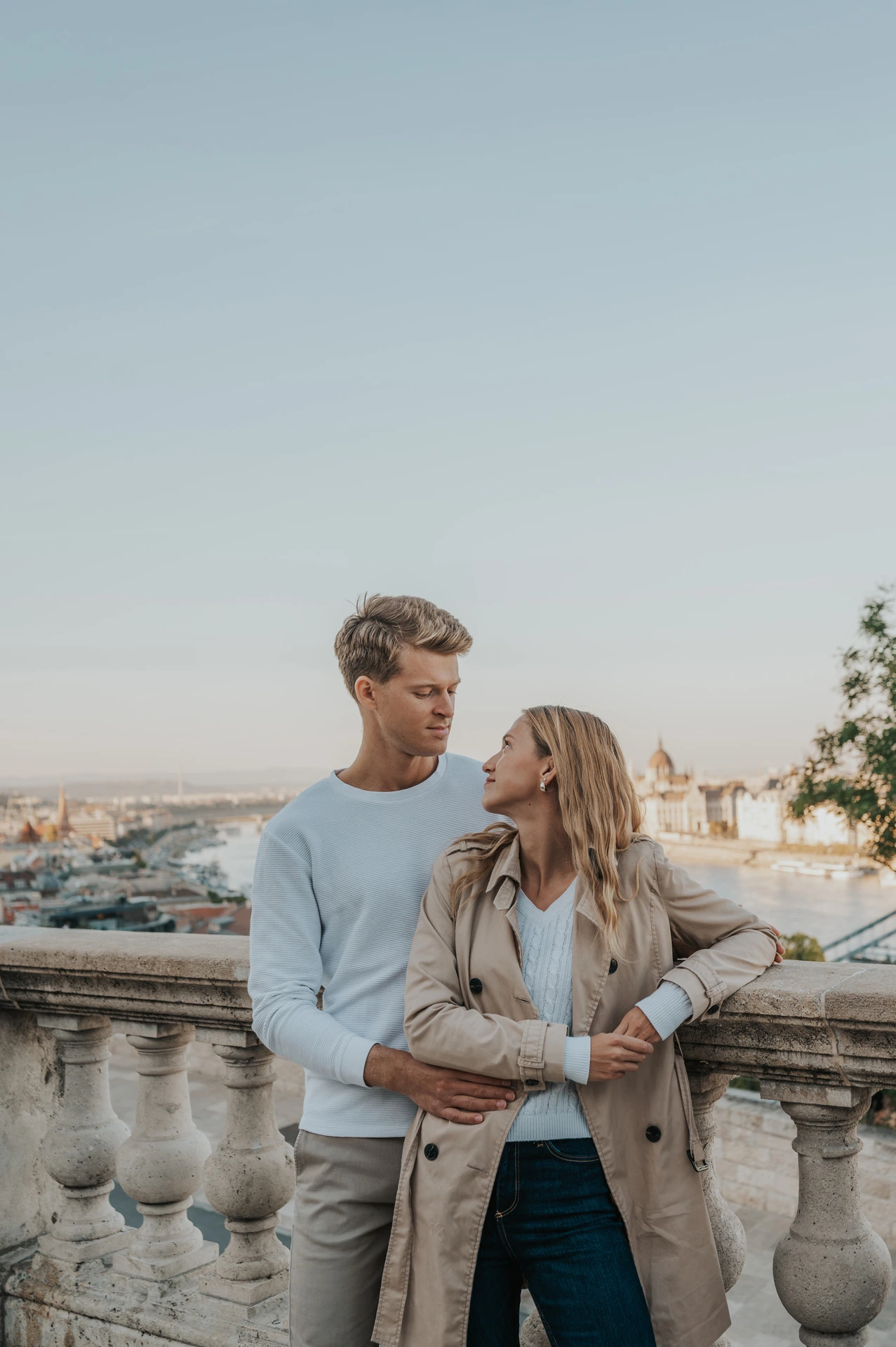 The couple embracing beside a stone balustrade with Budapest’s rooftops beyond Buda Castle during their couple photoshoot in Budapest.