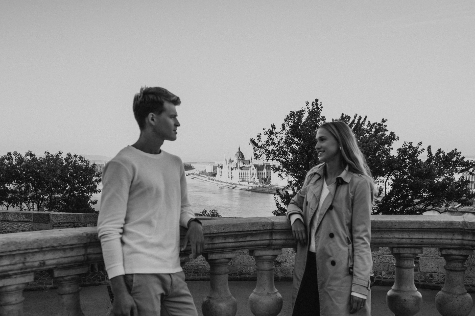 Black-and-white landscape portrait of the couple standing together beside Buda Castle’s stone balustrade during their couple photoshoot in Budapest.