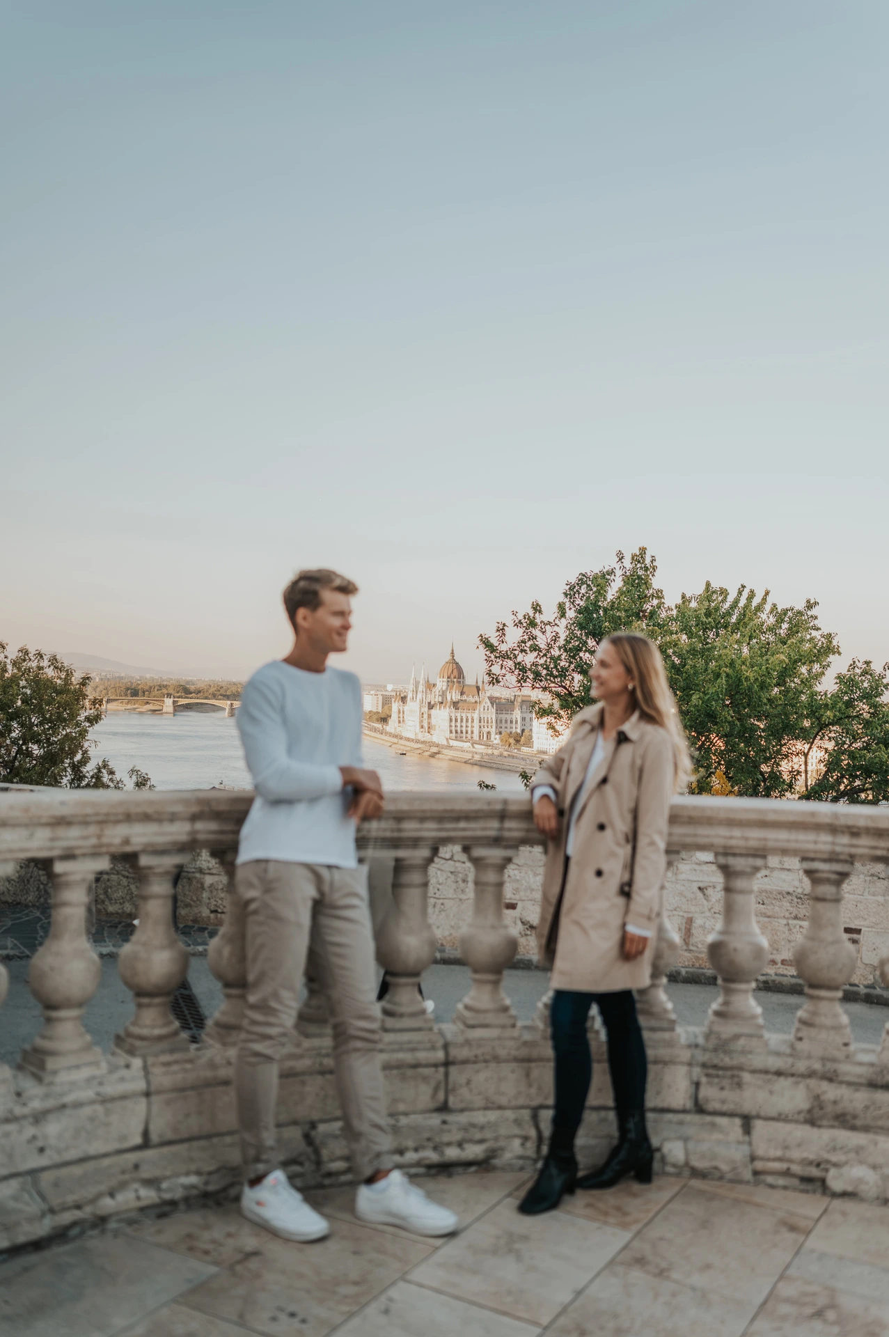 A full-length portrait of the couple standing beside a stone balustrade overlooking Budapest from Buda Castle during their couple photoshoot in Budapest.
