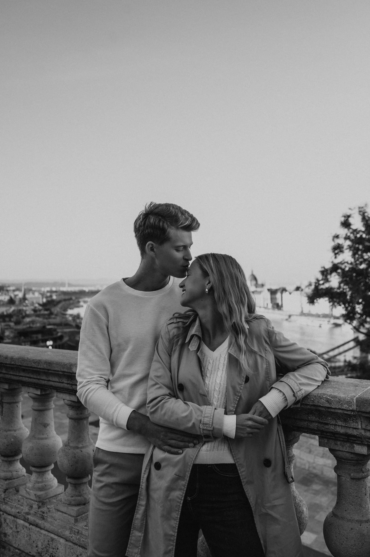 Black-and-white portrait of the couple embracing beside a stone balustrade overlooking Budapest from Buda Castle during their couple photoshoot in Budapest.