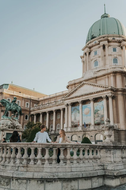 The couple beside a stone balustrade with Buda Castle’s green dome and pale palace façade rising behind them during their couple photoshoot in Budapest.