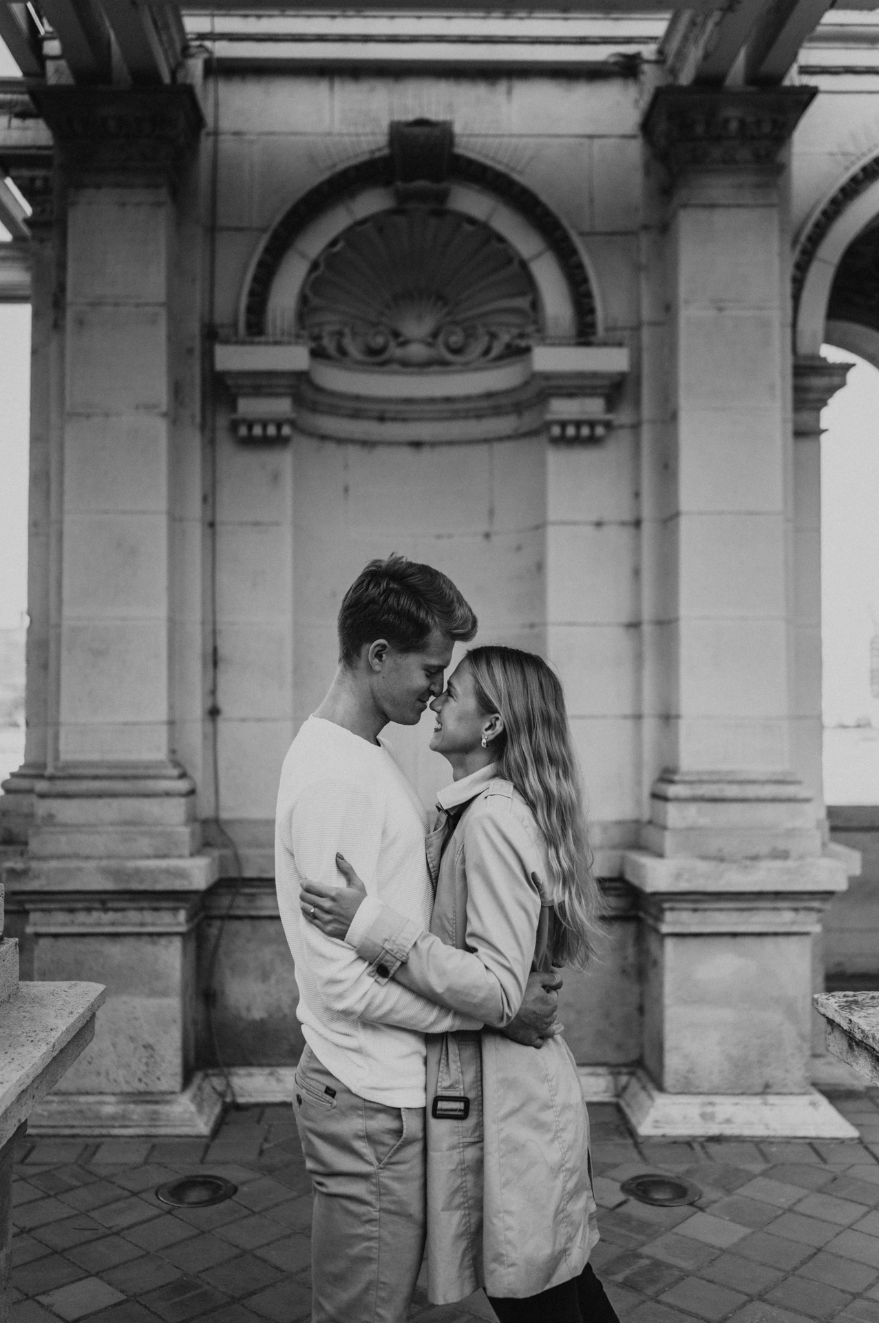 Black-and-white portrait of the couple embracing beneath Castle Garden Bazaar’s carved pavilion arch during their couple photoshoot in Budapest.