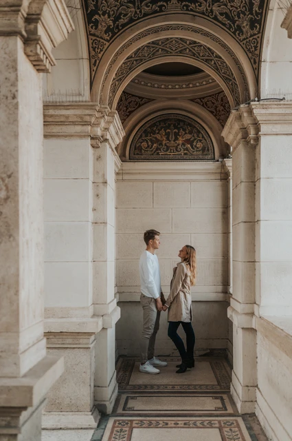 A walk through the ornate garden arches at a Budapest couple photoshoot The couple walking hand in hand through an ornate arched passage at Castle Garden Bazaar during their couple photoshoot in Budapest.