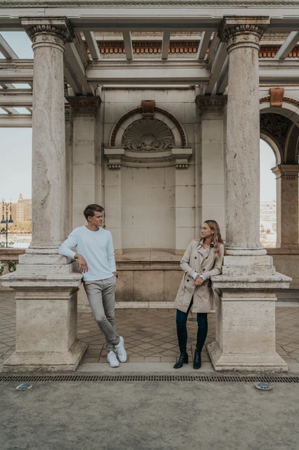 Time together between the pavilion columns at a Budapest couple photoshoot A wide portrait of the couple looking at one another beside a stone ledge between Castle Garden Bazaar’s pavilion columns during their couple photoshoot in Budapest.
