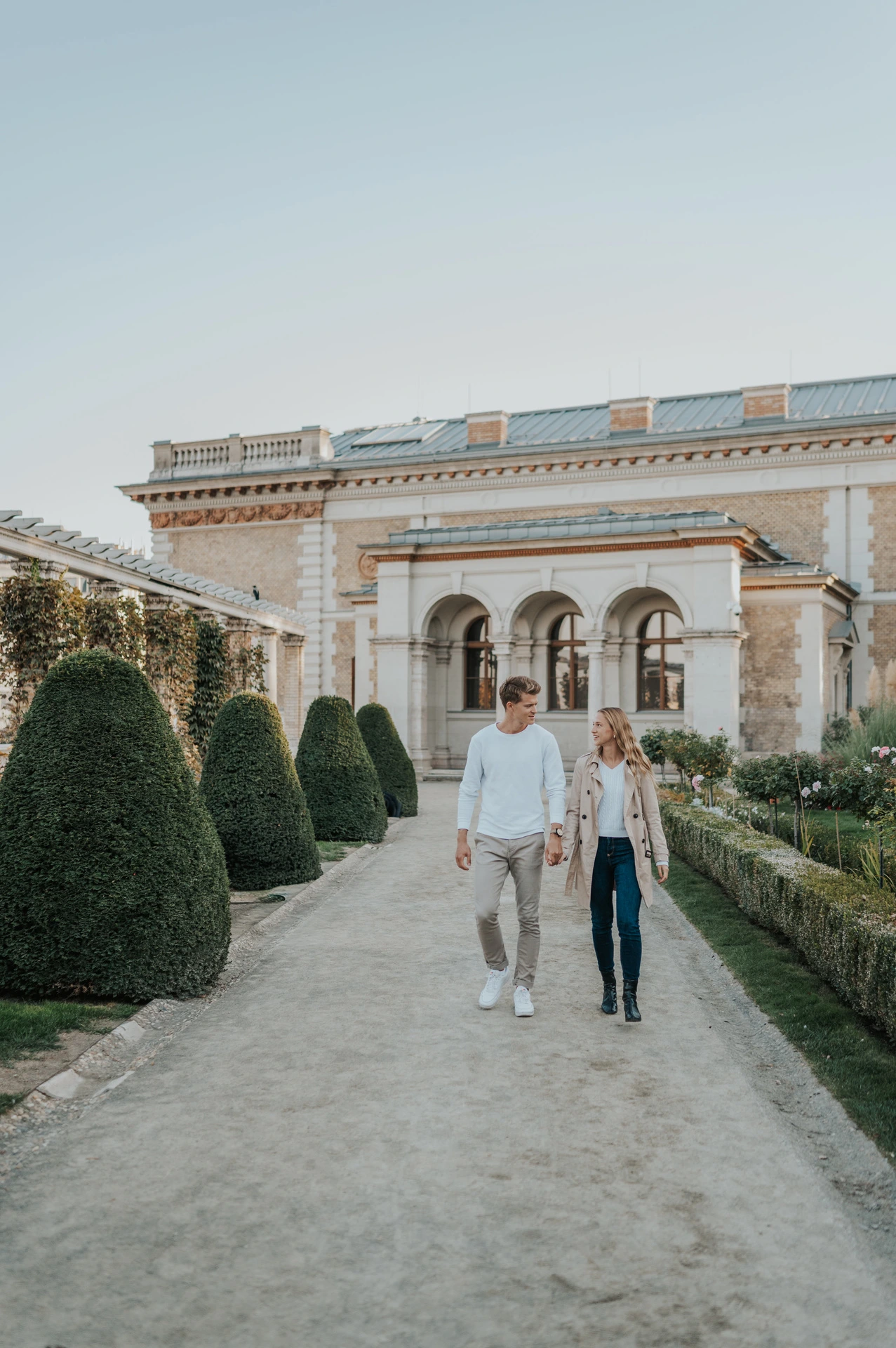 The couple walking hand in hand along a gravel path between clipped hedges at Castle Garden Bazaar during their couple photoshoot in Budapest.