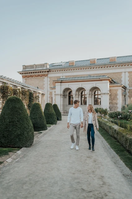 The couple walking hand in hand along a gravel path between clipped hedges at Castle Garden Bazaar during their couple photoshoot in Budapest.