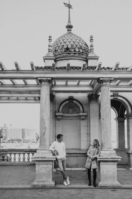 A pavilion portrait in black and white at a Budapest couple photoshoot Black-and-white portrait of the couple beside a stone ledge beneath Castle Garden Bazaar’s ornate pavilion and green roof during their couple photoshoot in Budapest.