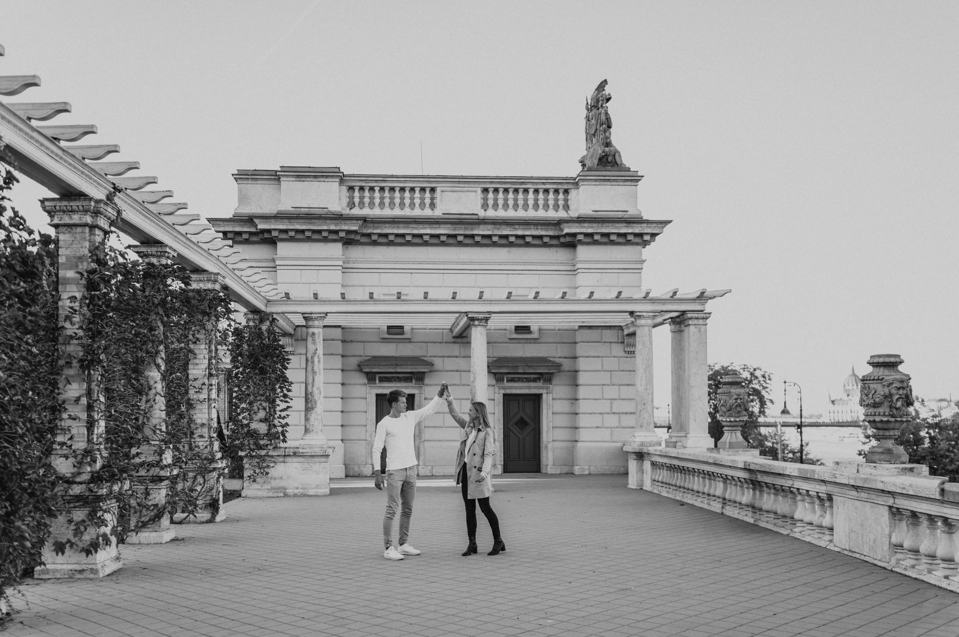 Black-and-white landscape portrait of the couple raising their joined hands beneath the pergola beside Castle Garden Bazaar’s pavilion during their couple photoshoot in Budapest.