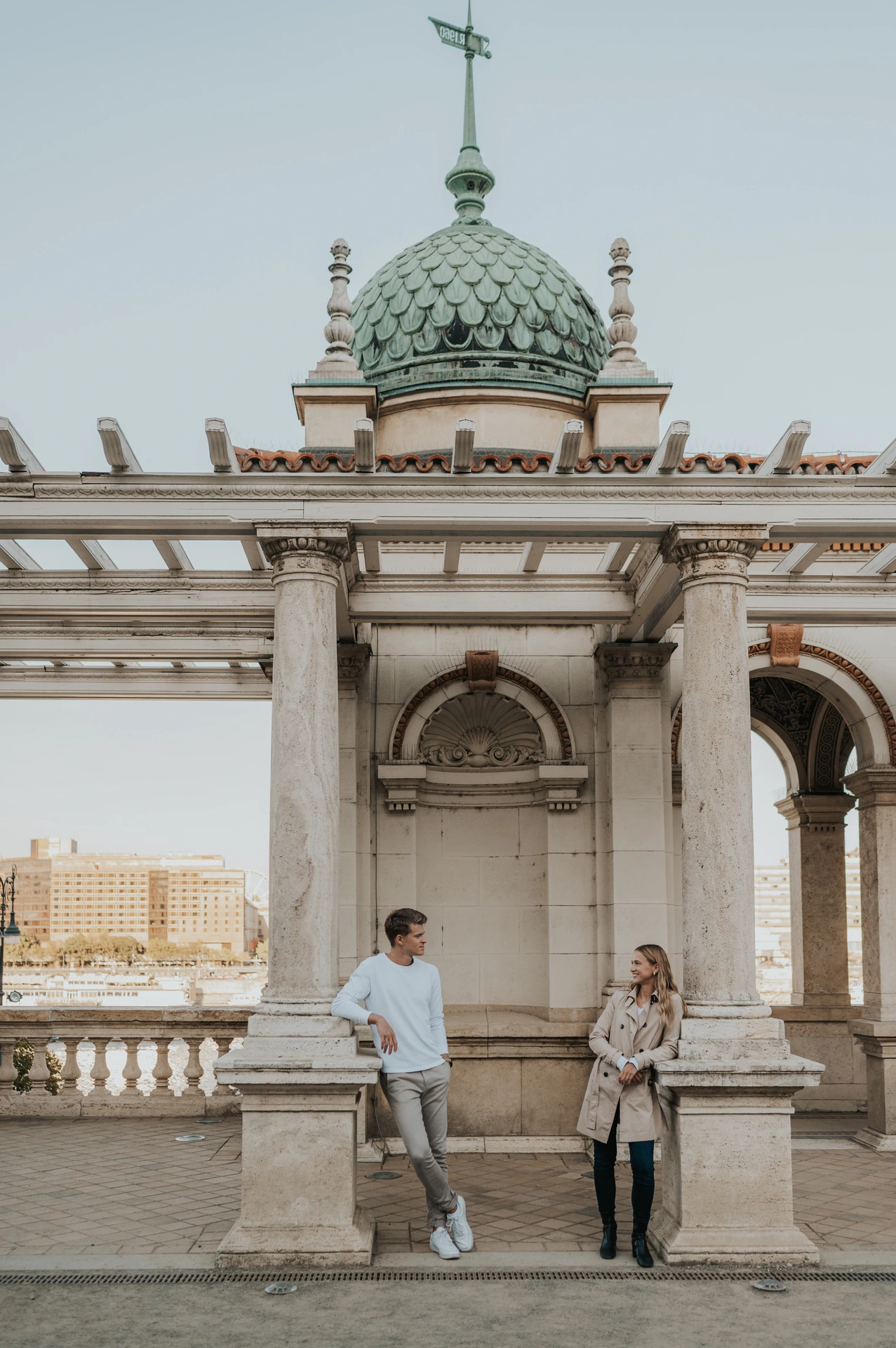 The couple standing beside a stone ledge beneath Castle Garden Bazaar’s ornate pavilion and green roof during their couple photoshoot in Budapest.