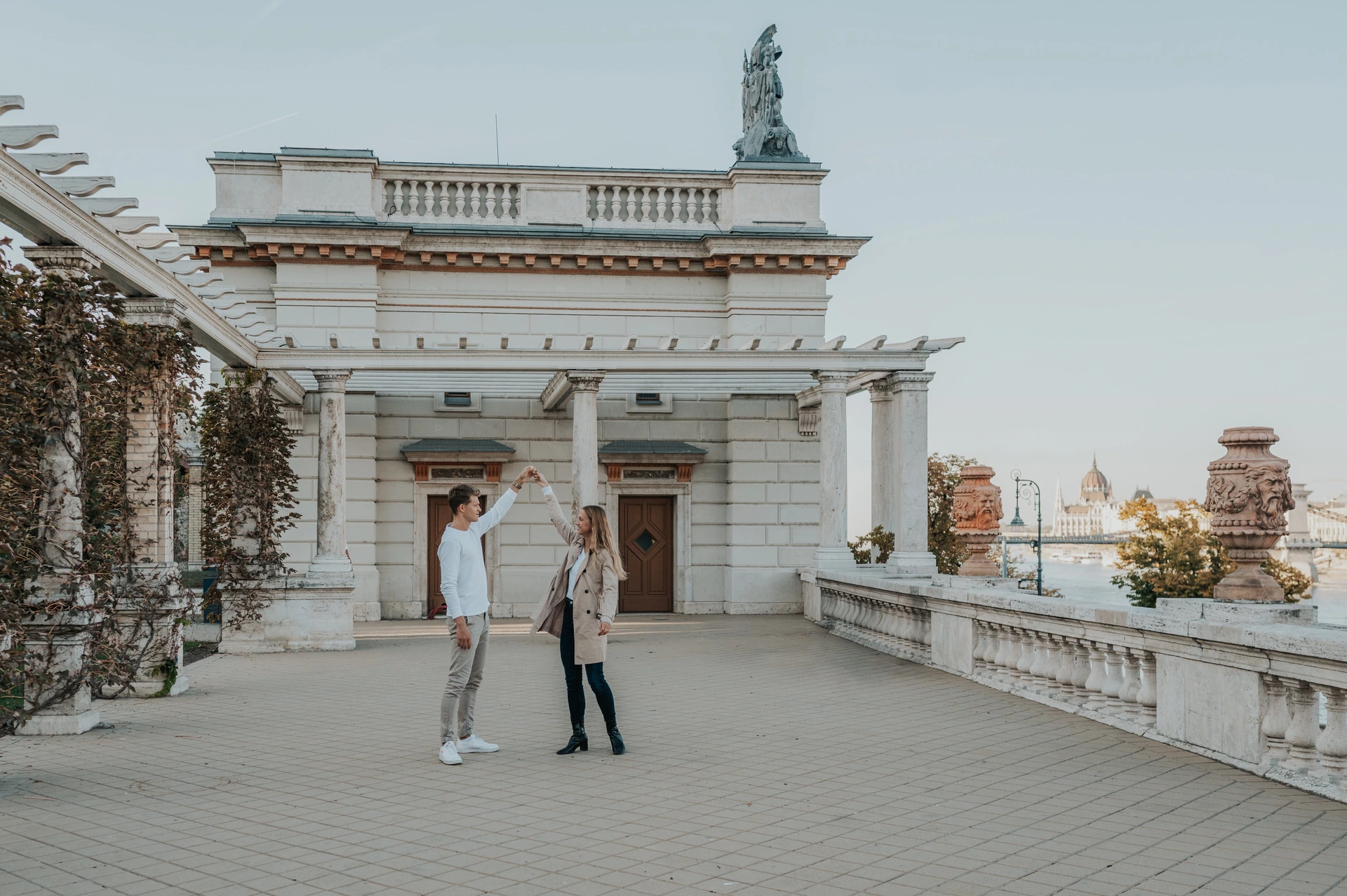 A landscape portrait of the couple raising their joined hands on the terrace beside Castle Garden Bazaar’s pale pavilion during their couple photoshoot in Budapest.