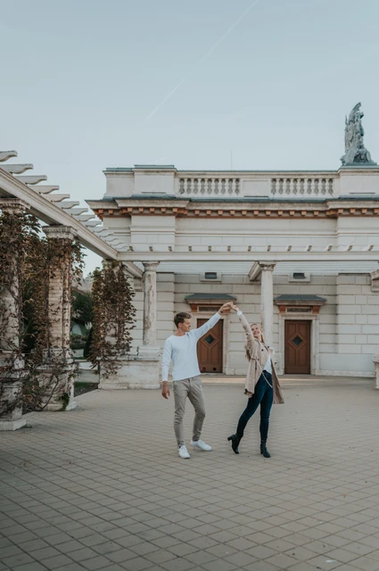 A turn beneath the blue Budapest sky at a Budapest couple photoshoot The couple raising their joined hands beside Castle Garden Bazaar’s pavilion beneath a blue sky during their couple photoshoot in Budapest.