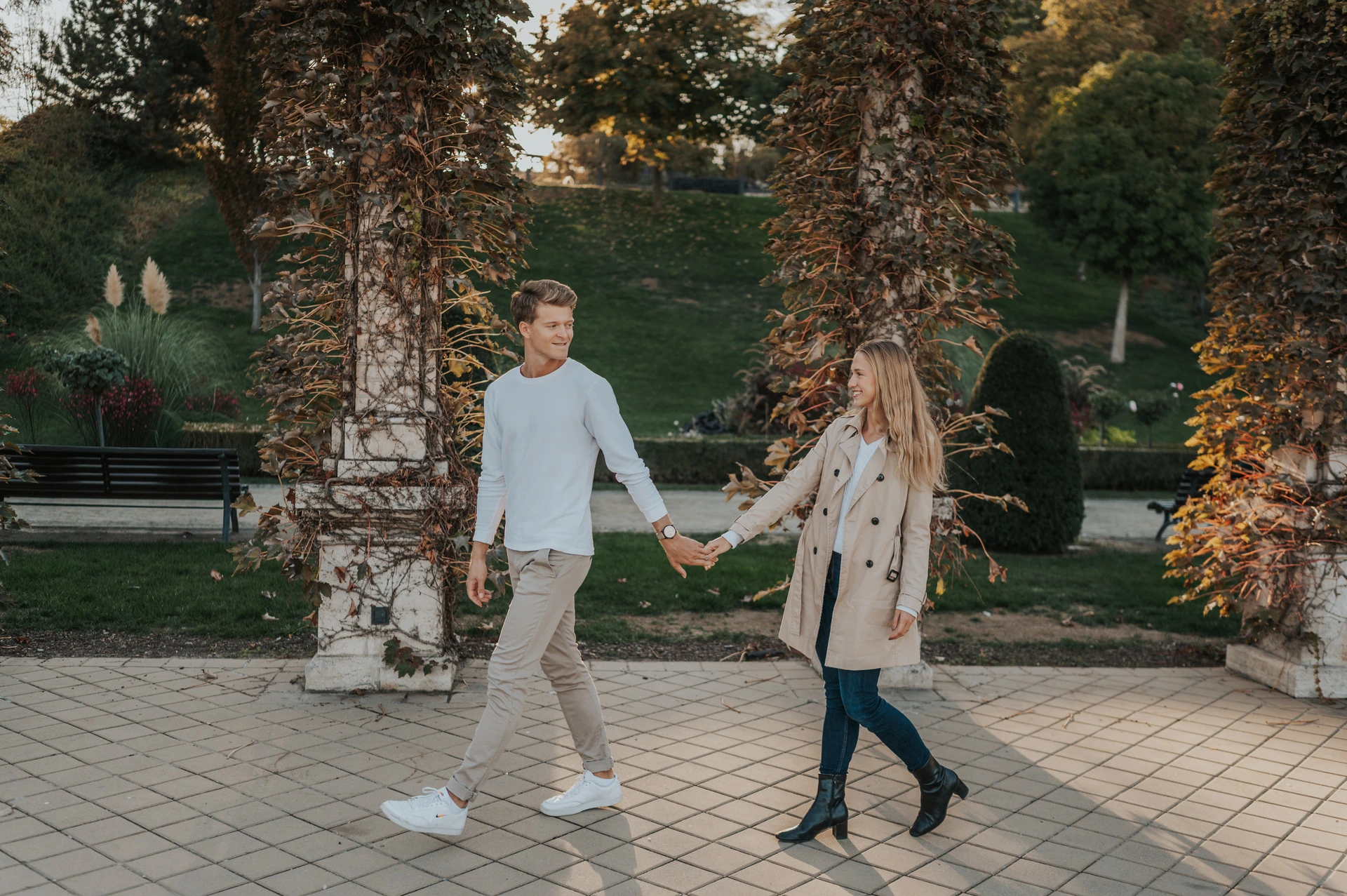 A landscape portrait of the couple walking hand in hand between leafy pergola columns at Castle Garden Bazaar during their couple photoshoot in Budapest.