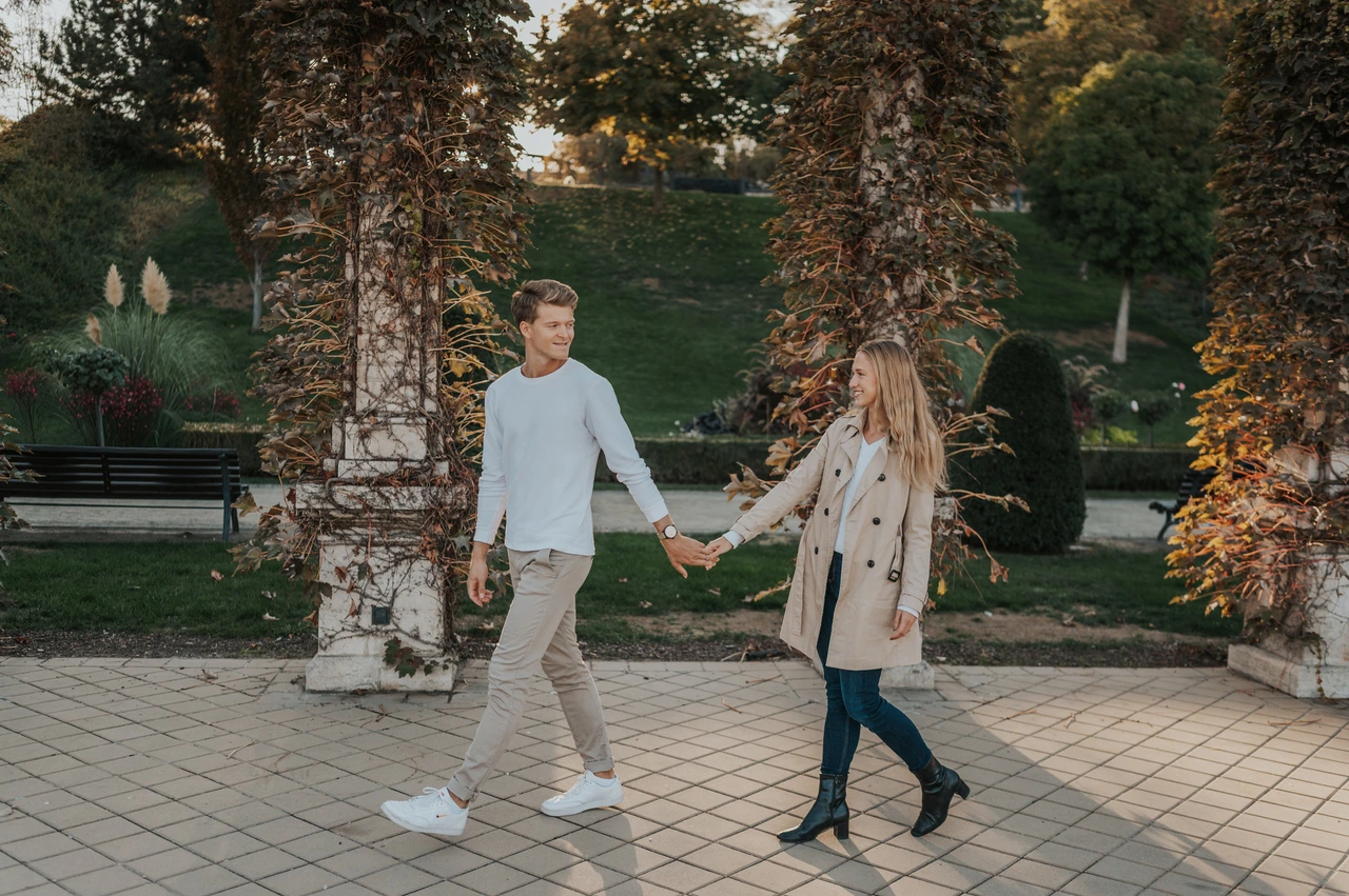 A wide view beneath the garden pergola at a Budapest couple photoshoot A landscape portrait of the couple walking hand in hand between leafy pergola columns at Castle Garden Bazaar during their couple photoshoot in Budapest.