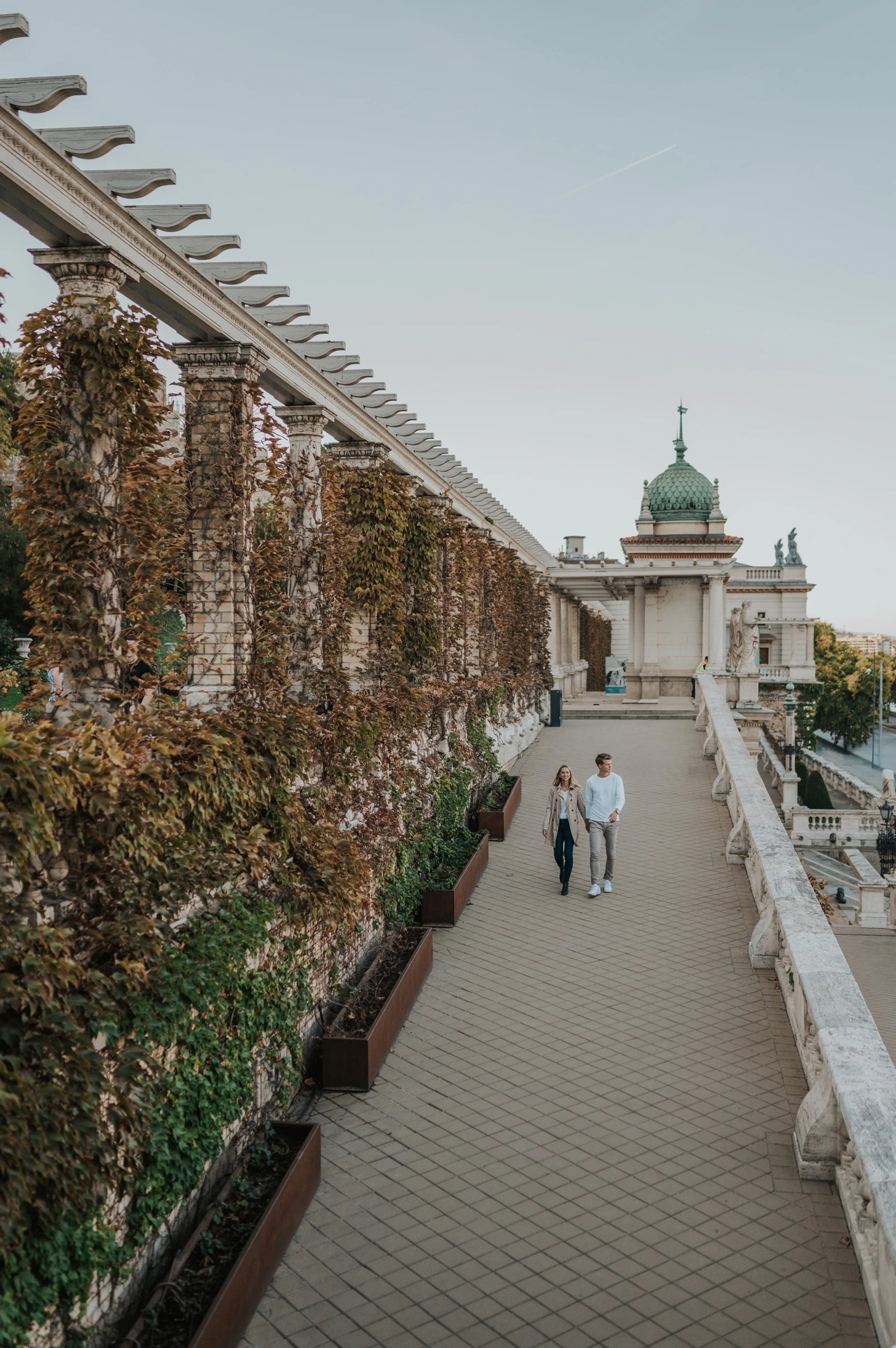 A wide view of the couple walking down the terrace steps beside Castle Garden Bazaar’s long ivy-covered pergola during their couple photoshoot in Budapest.