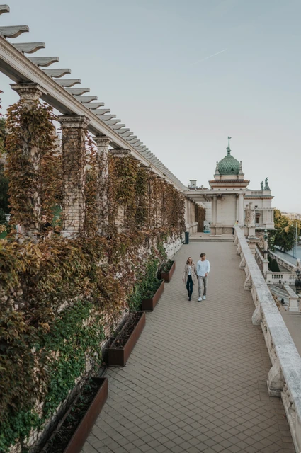 A walk beside the long garden pergola at a Budapest couple photoshoot A wide view of the couple walking down the terrace steps beside Castle Garden Bazaar’s long ivy-covered pergola during their couple photoshoot in Budapest.