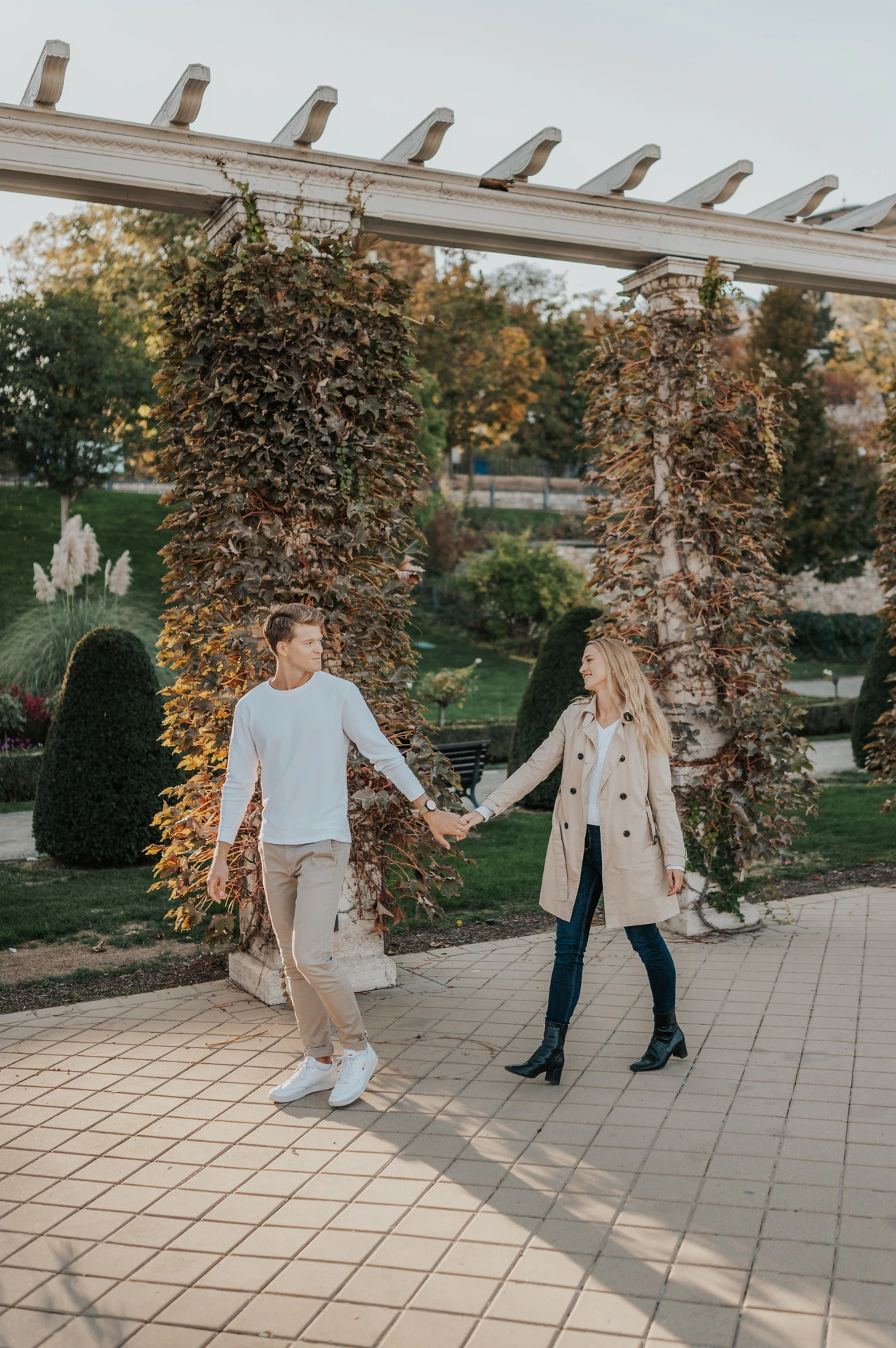The couple walking hand in hand beneath leafy pergola columns at Castle Garden Bazaar during their couple photoshoot in Budapest.