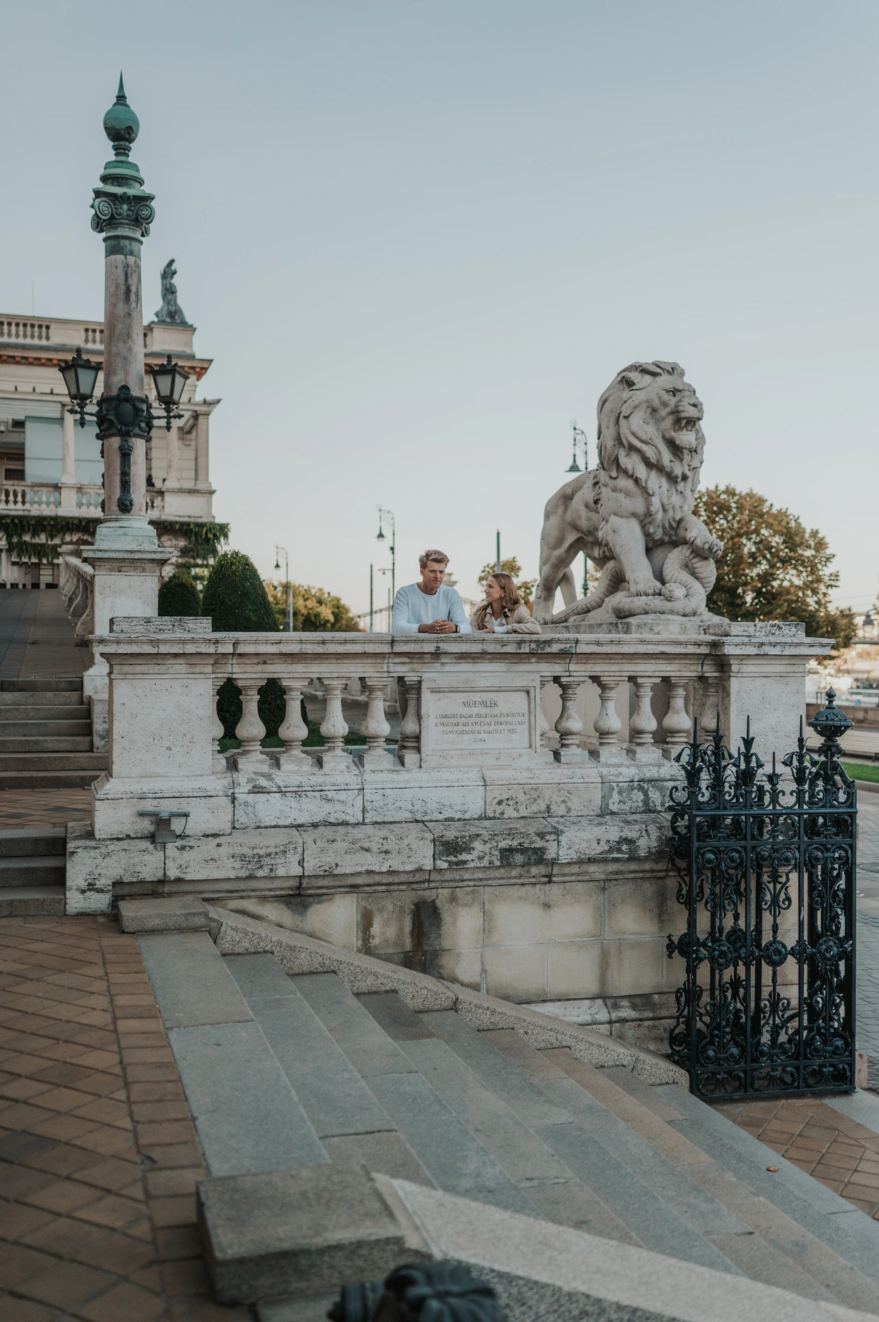 A wide portrait of the couple behind a stone balustrade beside a large lion statue and ornate green lamp at Castle Garden Bazaar during their couple photoshoot in Budapest.